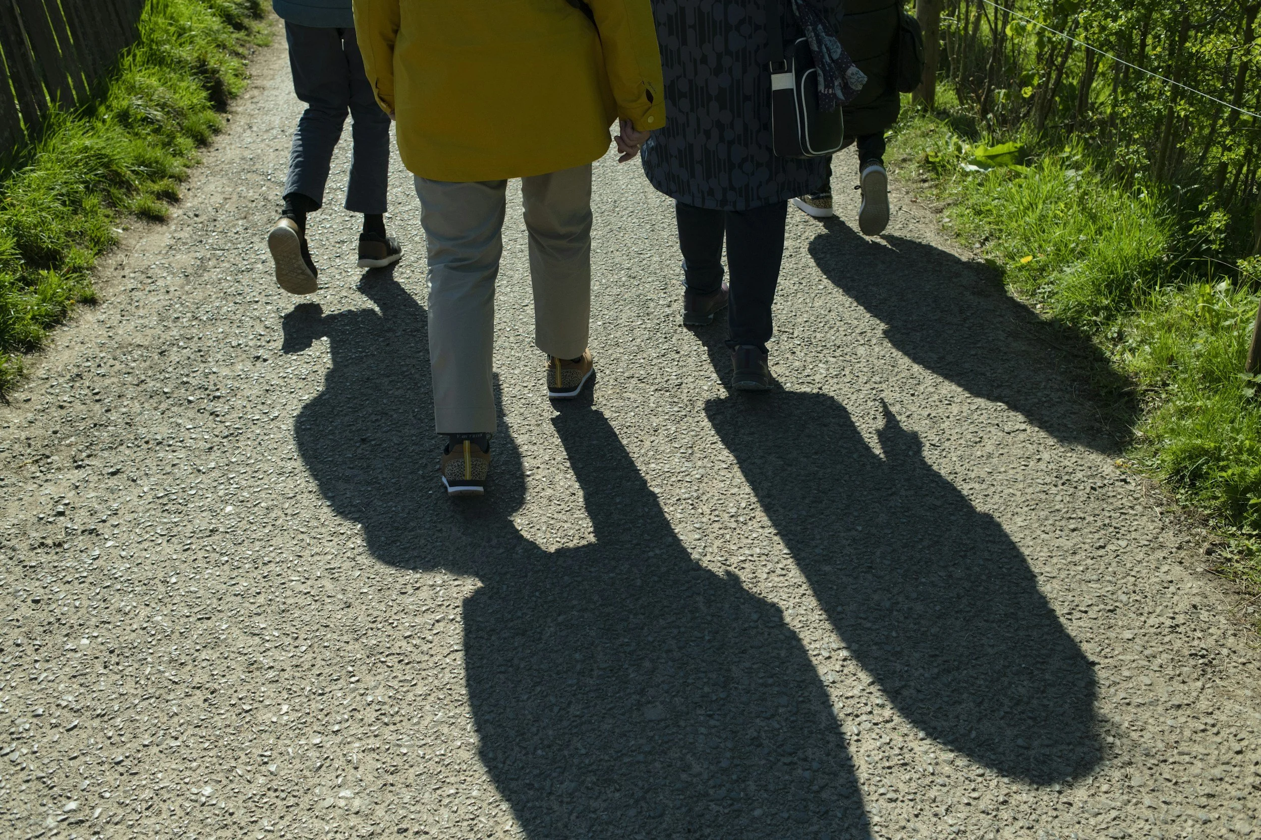 People walking in a park on a gravel path, casting long shadows, surrounded by greenery. (Faith Walk)