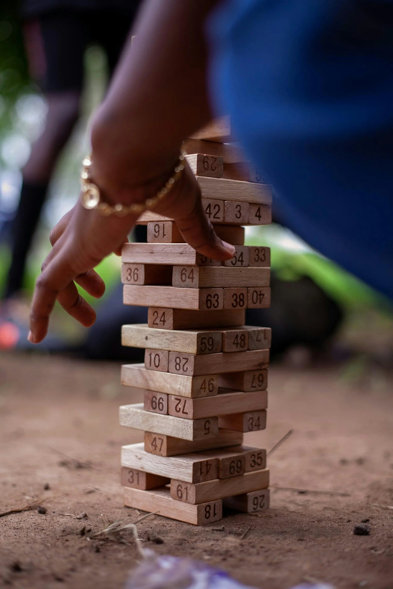 Person playing giant Jenga game outdoors on dirt ground, wearing a blue shirt and gold bracelet. (Spark Game Nights)