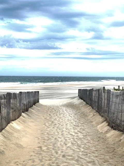 Modern building with wooden exterior surrounded by grassy plants, mountains in the background, and two people visible through a large glass window.