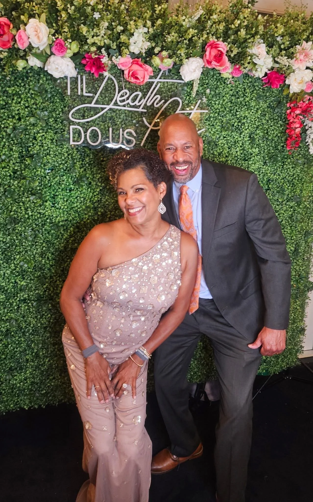 Smiling man and woman dressed in formal attire standing in front of a green floral wall with pink, white, and red flowers (Spark LJC Founders)