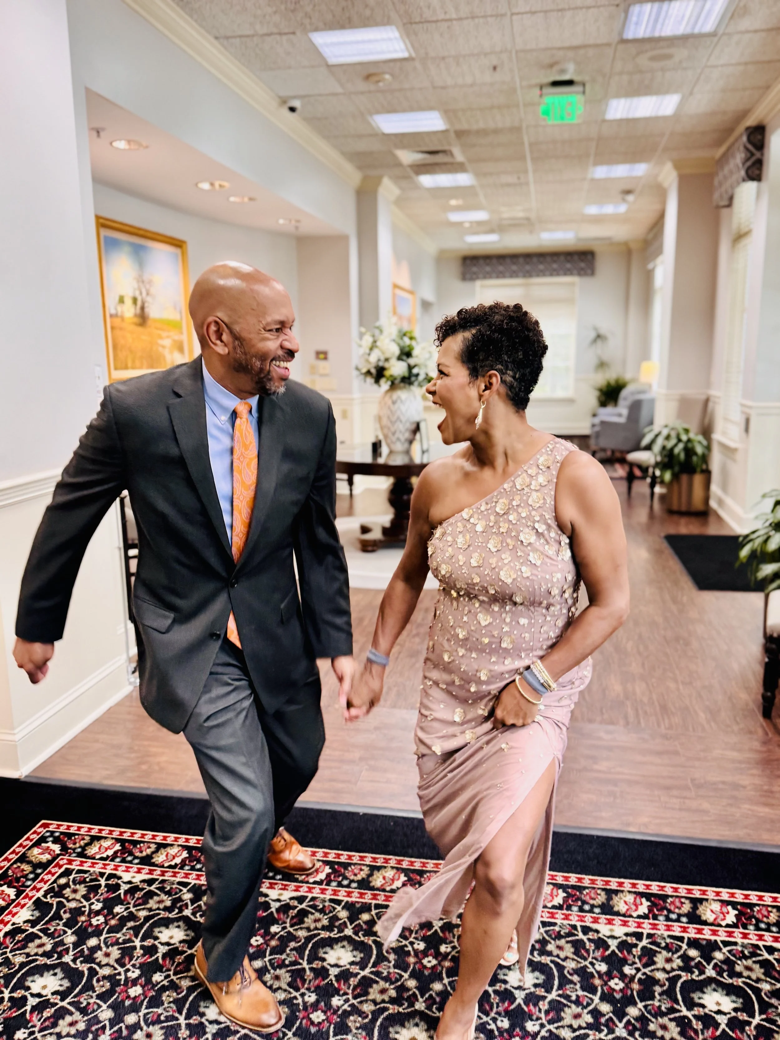 A man and woman holding hands and smiling in a decorated indoor setting with paintings, plants, and furniture.