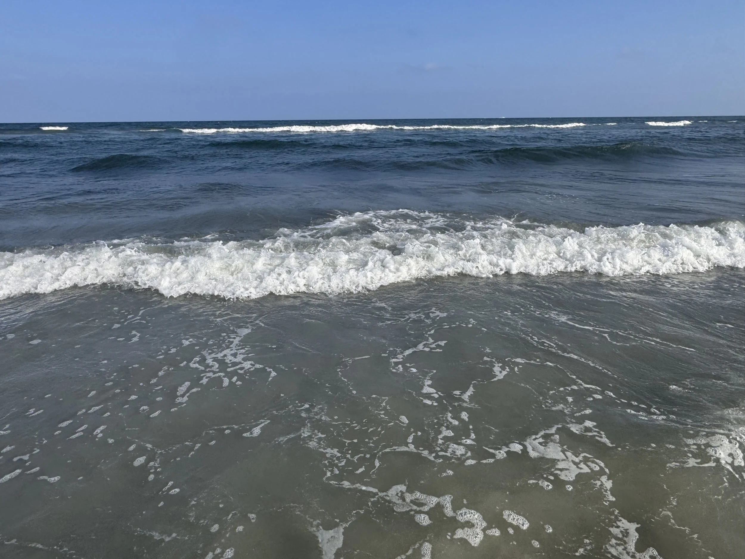 Ocean waves crashing on sandy beach with blue sky above.