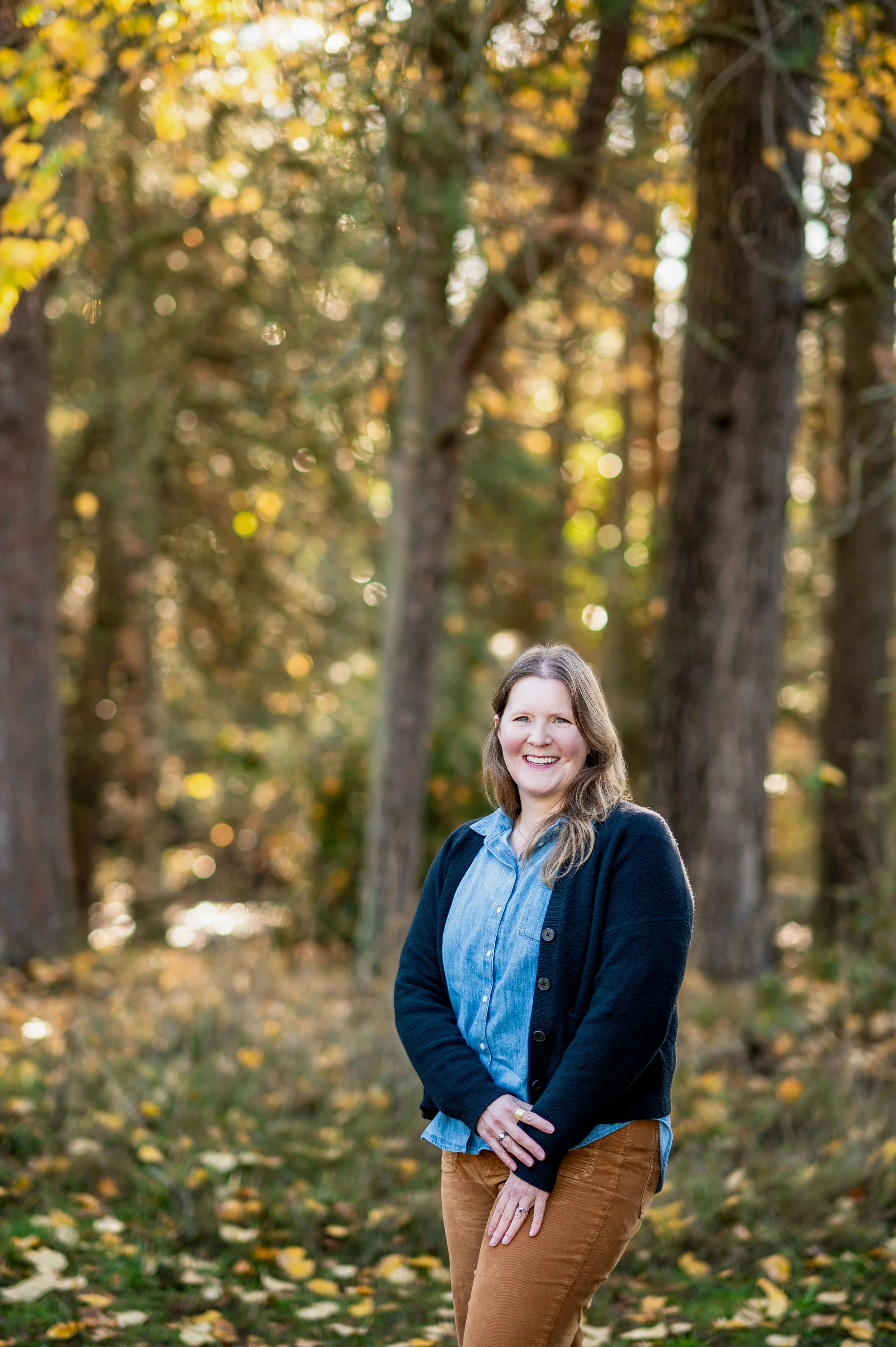 A woman standing outdoors in a forest during autumn, smiling, with trees and fallen leaves in the background.