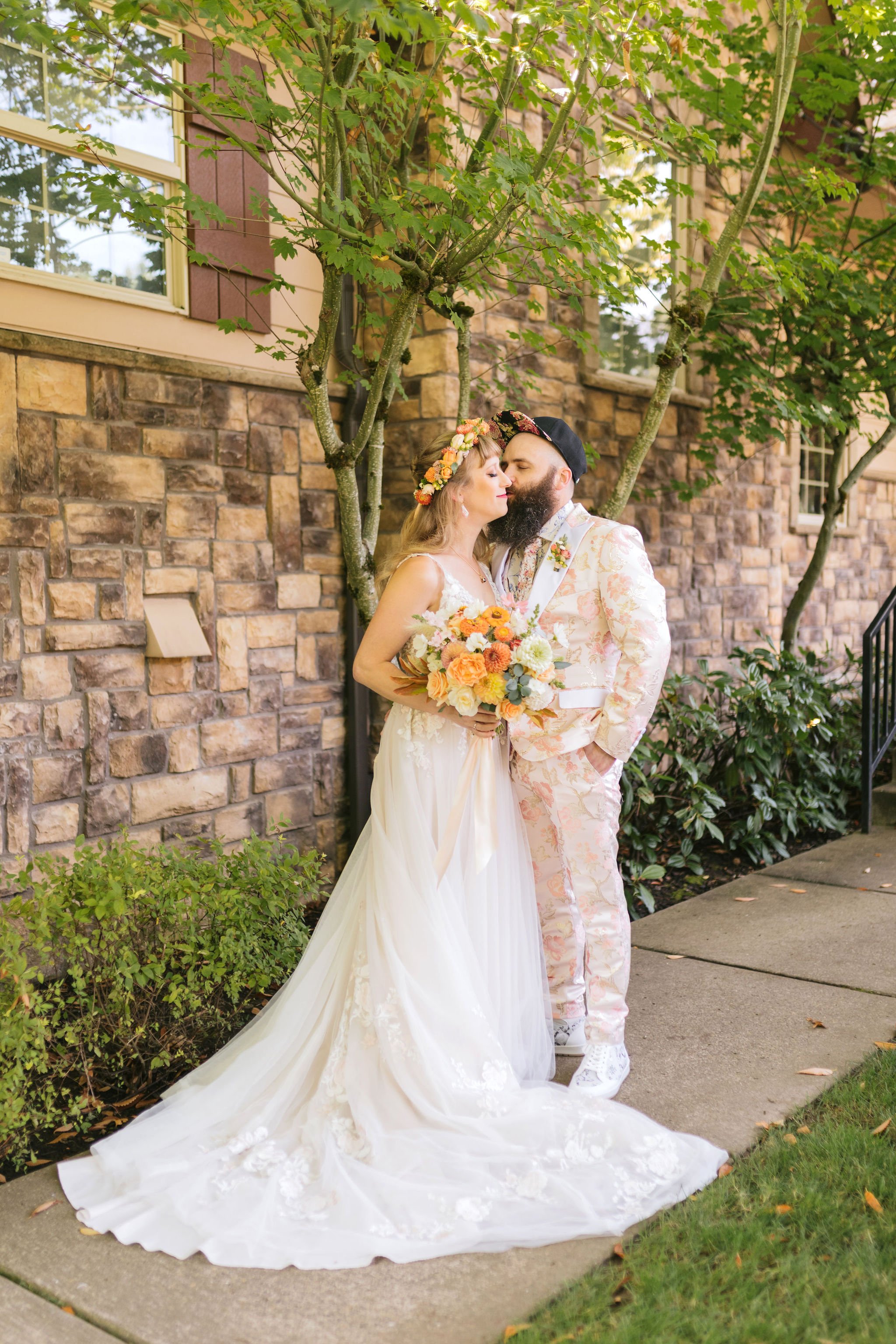A bride and groom stand close together outdoors, with the bride holding a colorful bouquet. The bride wears a white wedding dress with a train and a floral headband, and the groom wears a light pastel suit with a matching floral pattern. They are near a brick wall and green foliage, sharing an intimate moment.