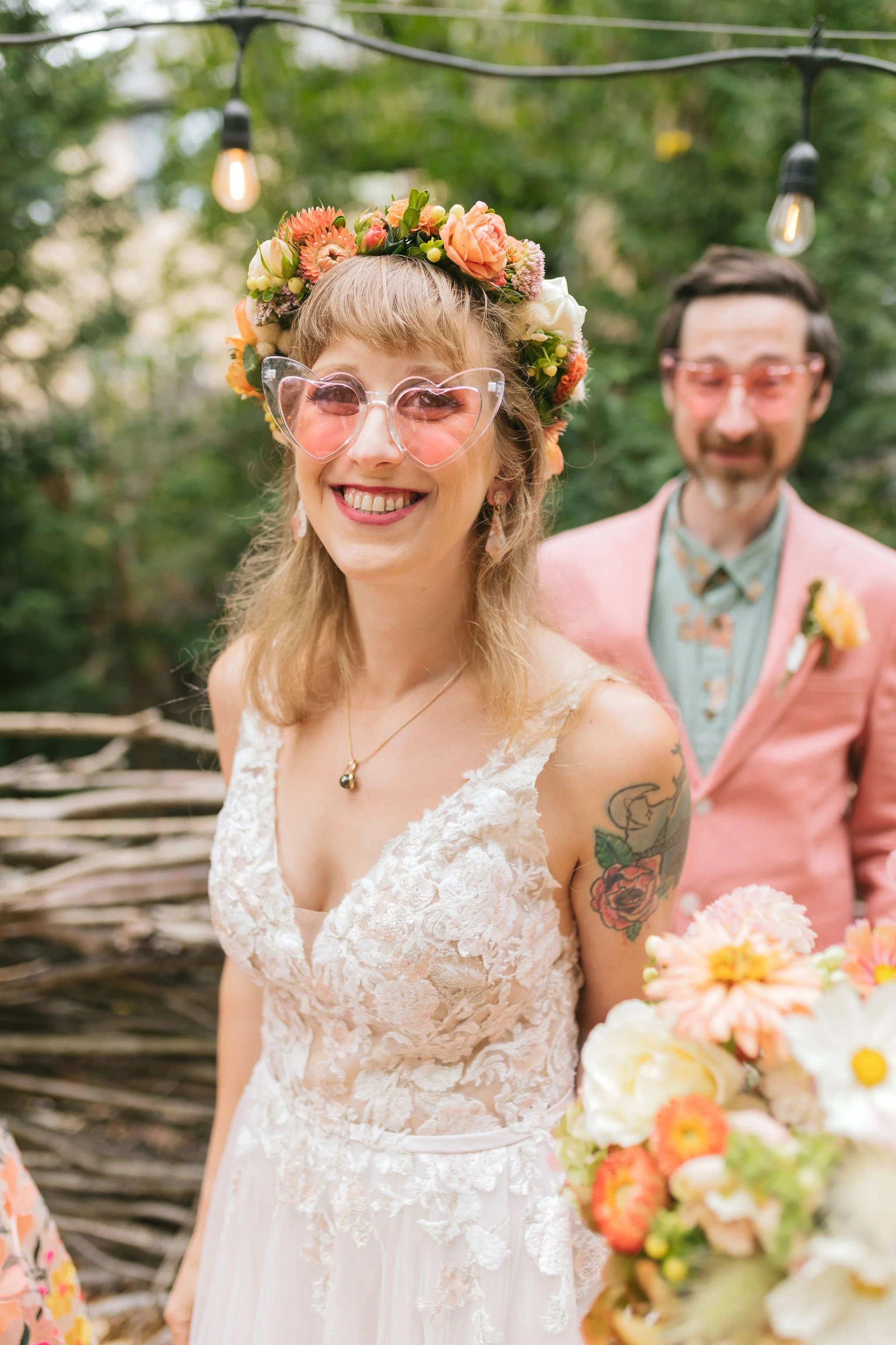 A smiling woman in a lace wedding dress wearing a floral crown and pink heart-shaped sunglasses, standing outdoors with a man in a pink suit and glasses behind her, surrounded by flowers and string lights in a lush green setting.

Photo by Char Marl 
