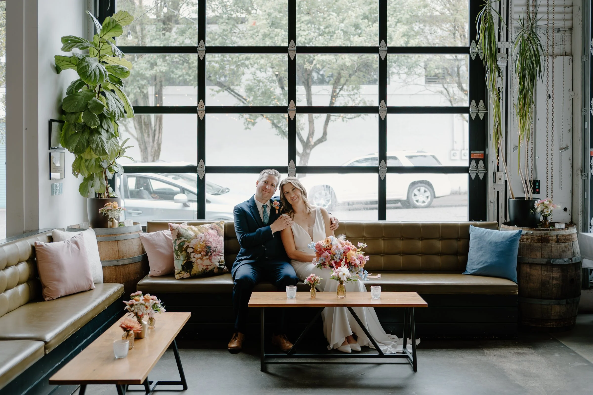 A couple in wedding attire sitting on a vintage sofa inside a venue with large windows. The man wears a dark blue suit and the woman a white wedding dress, both smiling and holding a bouquet of flowers. The venue has potted plants, colorful pillows, 