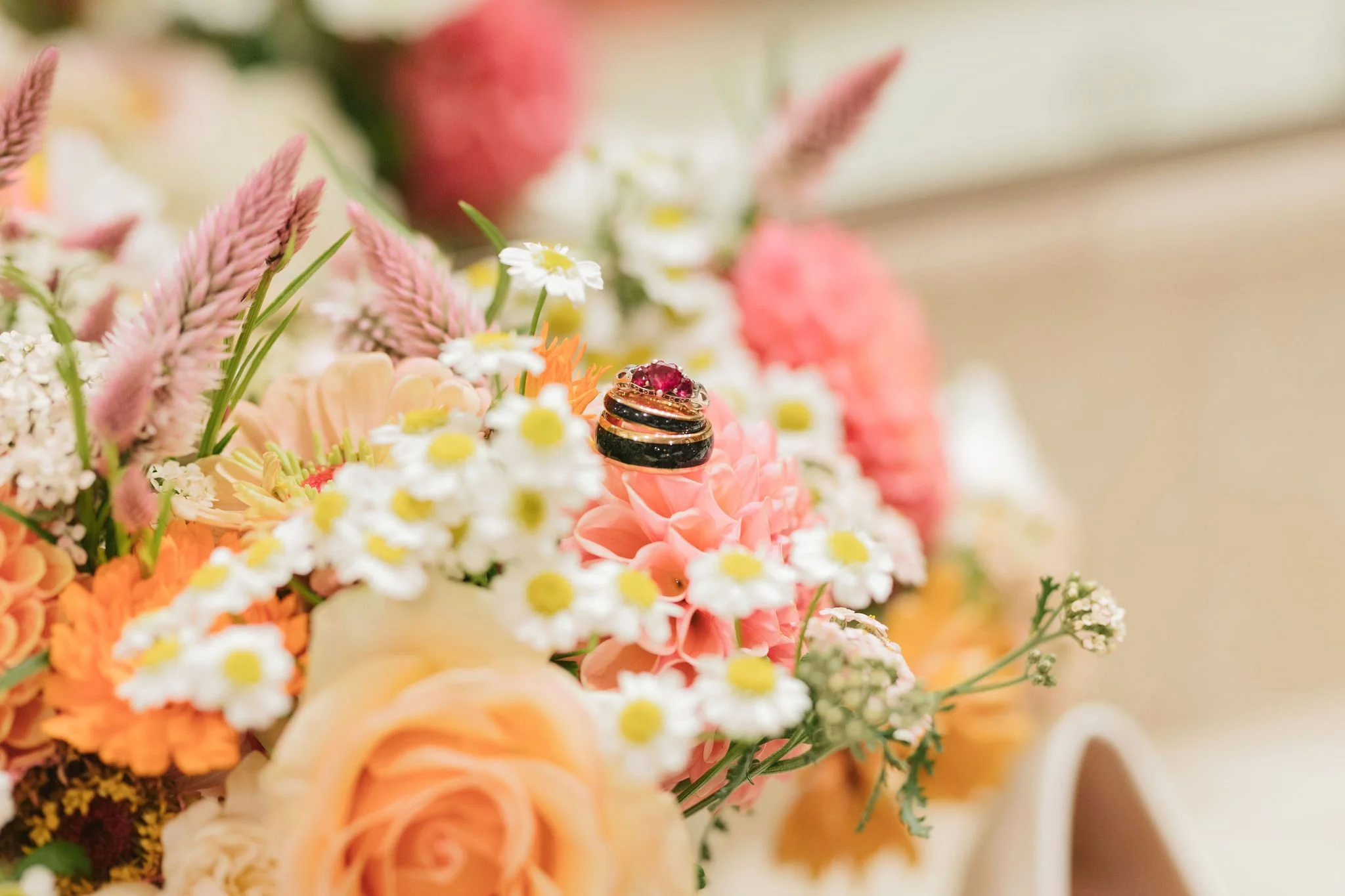 A close-up of a wedding bouquet with pink, white, and peach flowers, including daisies, roses, and other blooms, with wedding rings resting on a pink flower in the center.

Photo by Char Marl Photography
Venue: The Aerie at Eagles Landing in Happy Va