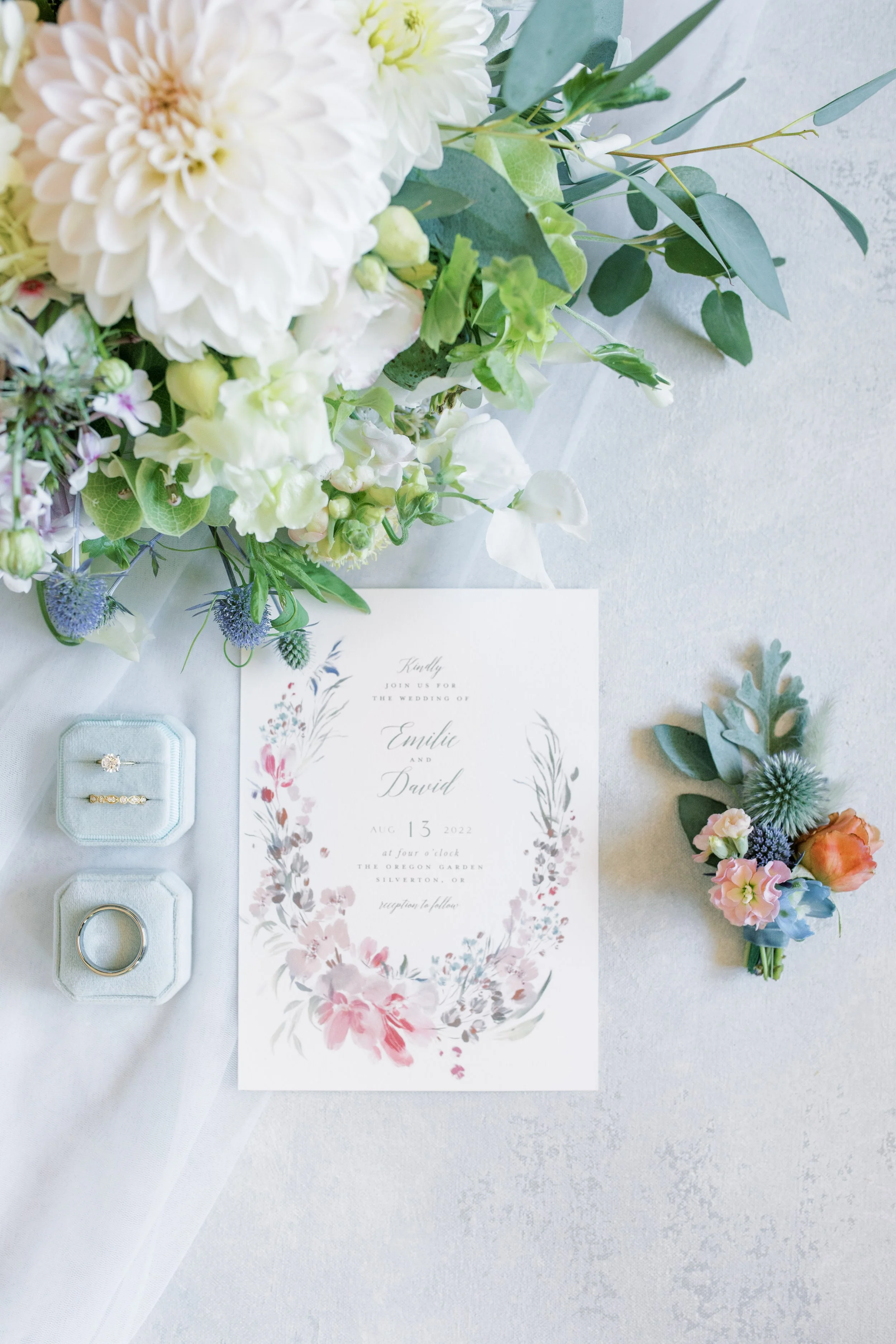 Wedding invitation with floral border, wedding rings in velvet boxes, and a boutonniere, surrounded by white and pastel pink, blue, and green flowers and foliage on a light background.

Photo by McKenna Rachelle Photography
Venue: The Oregon Gardens 