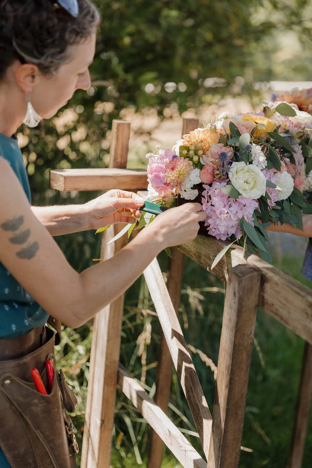 A woman is creating a flower arrangement outdoors on a wooden fence, surrounded by greenery on a sunny day.