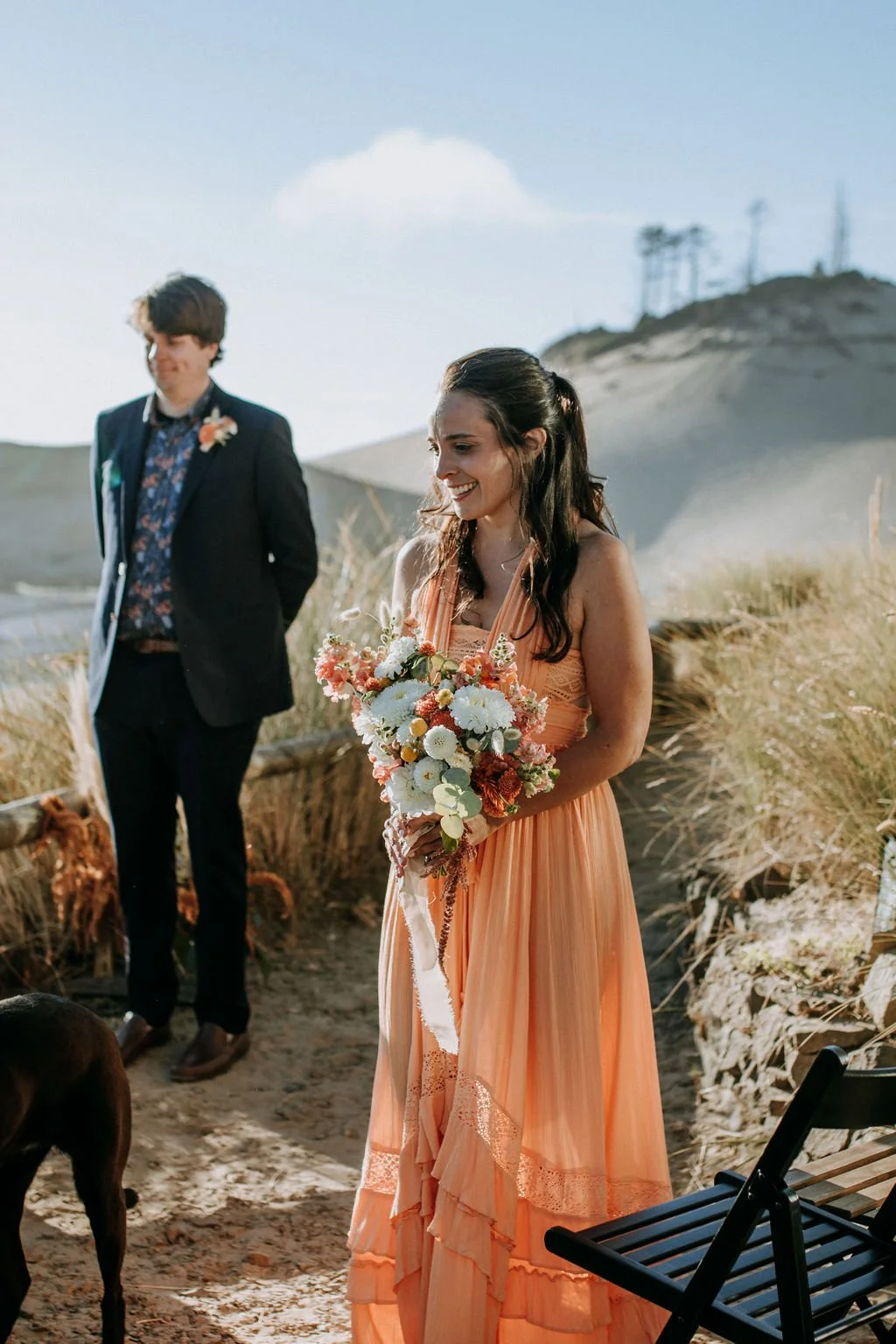 A woman in an orange dress holding a bouquet of flowers at a beach wedding, with a man in a suit standing in the background.