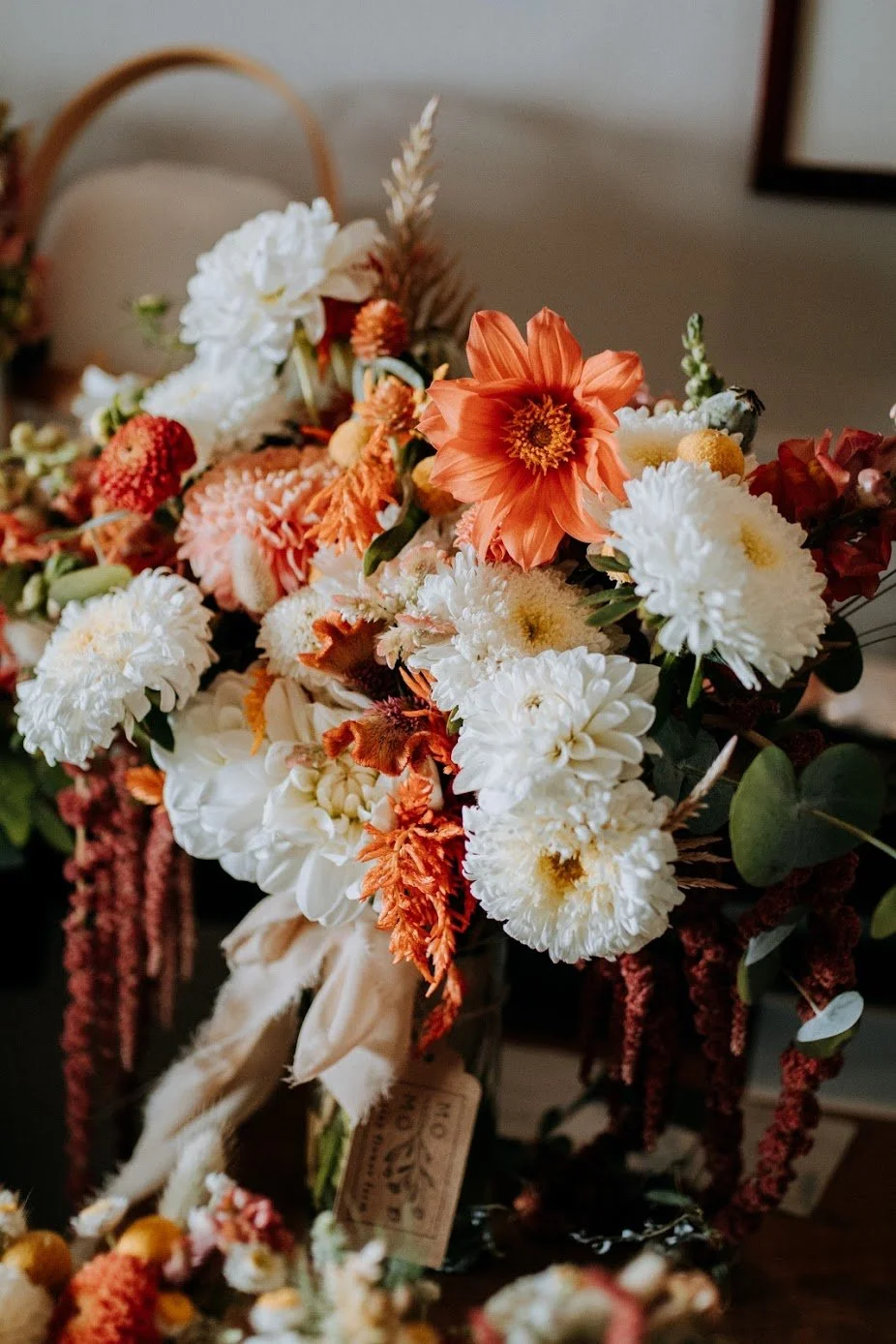 A vibrant bouquet of orange, white, and red flowers, including dahlias and other blooms, arranged in a rustic style with greenery, placed on a table indoors.