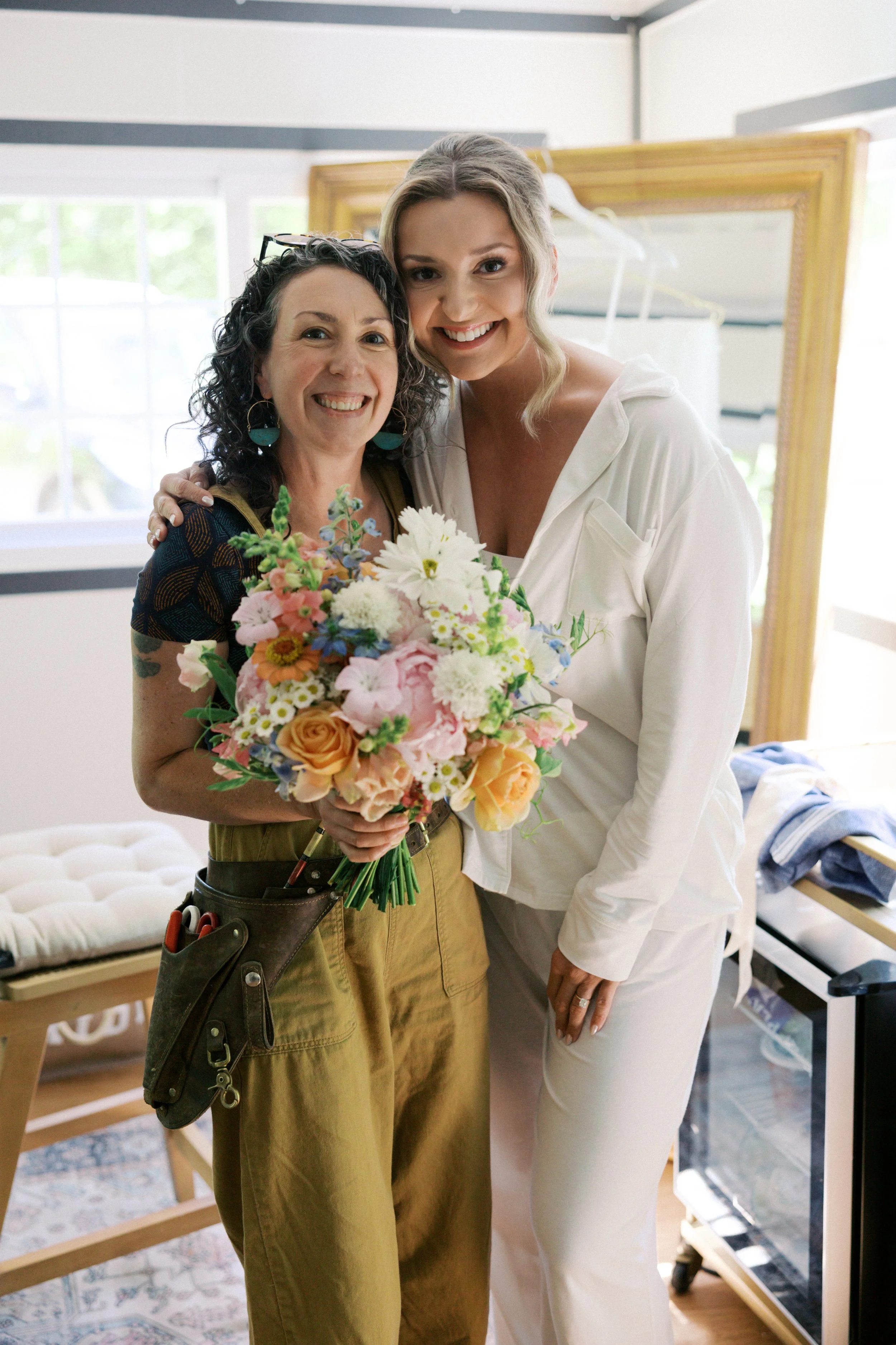 Two women smiling, one holding a bouquet of flowers, in a brightly lit room with a large mirror behind them. Photo by Here Today Photos