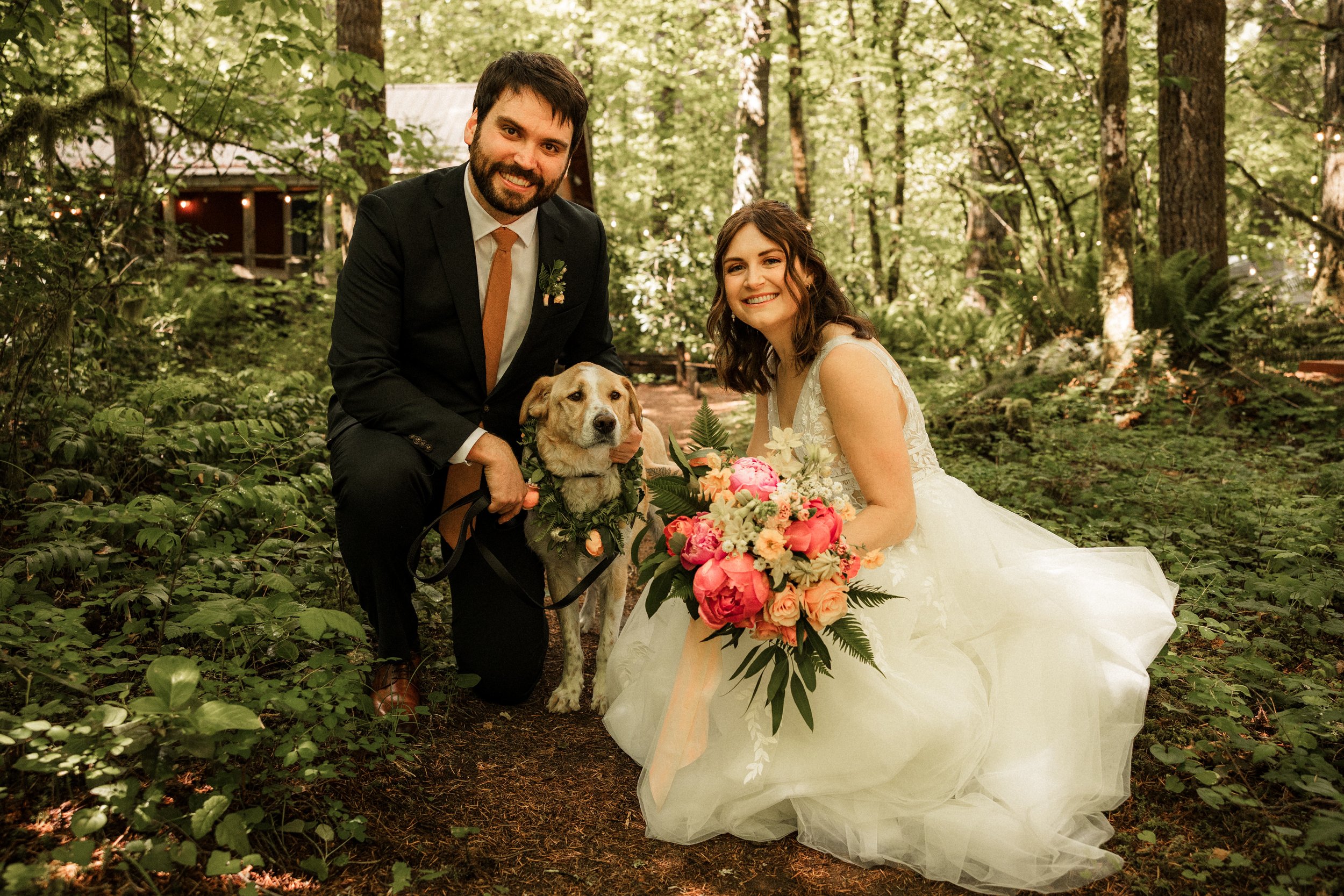 A newlywed couple with a dog in a forest. The groom in a black suit and tie, the bride in a white wedding dress holding a bouquet. The dog wears a flower collar. The background features trees and greenery. Photos by Works by Sarah Jane.