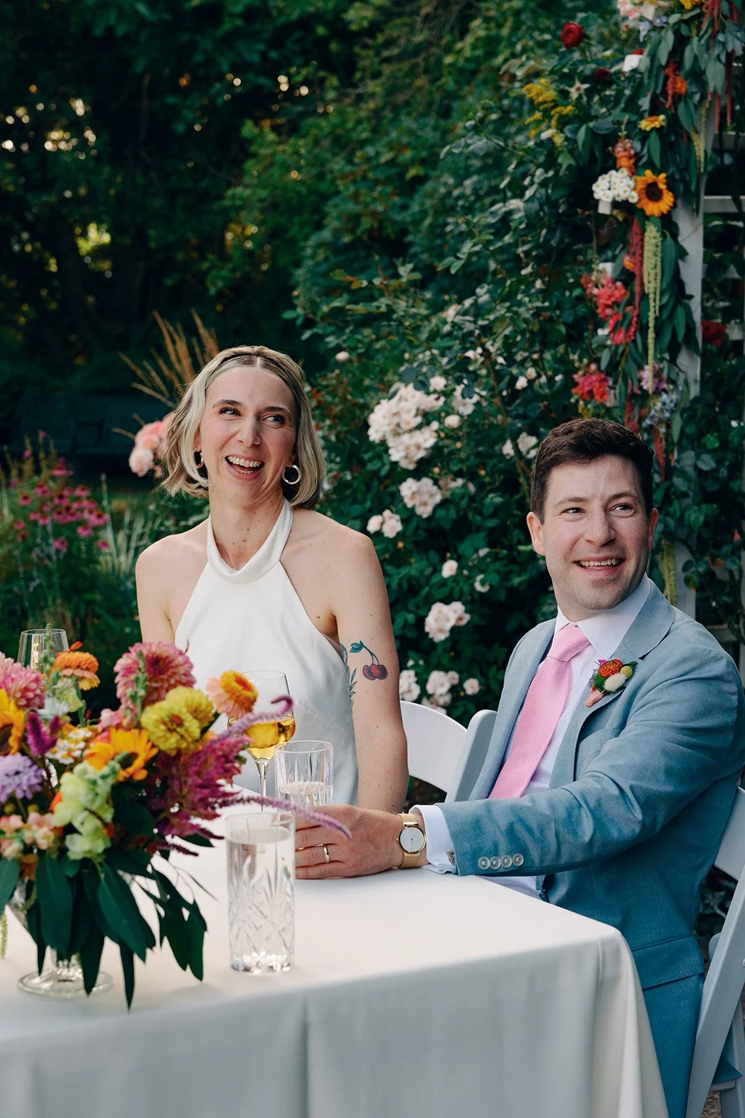 A woman and a man sitting at a table outdoors, smiling and laughing. The woman has blonde hair, wearing a white sleeveless dress, and the man has dark hair, wearing a light grey suit with a pink tie. There are flowers on the table and in the backgrou