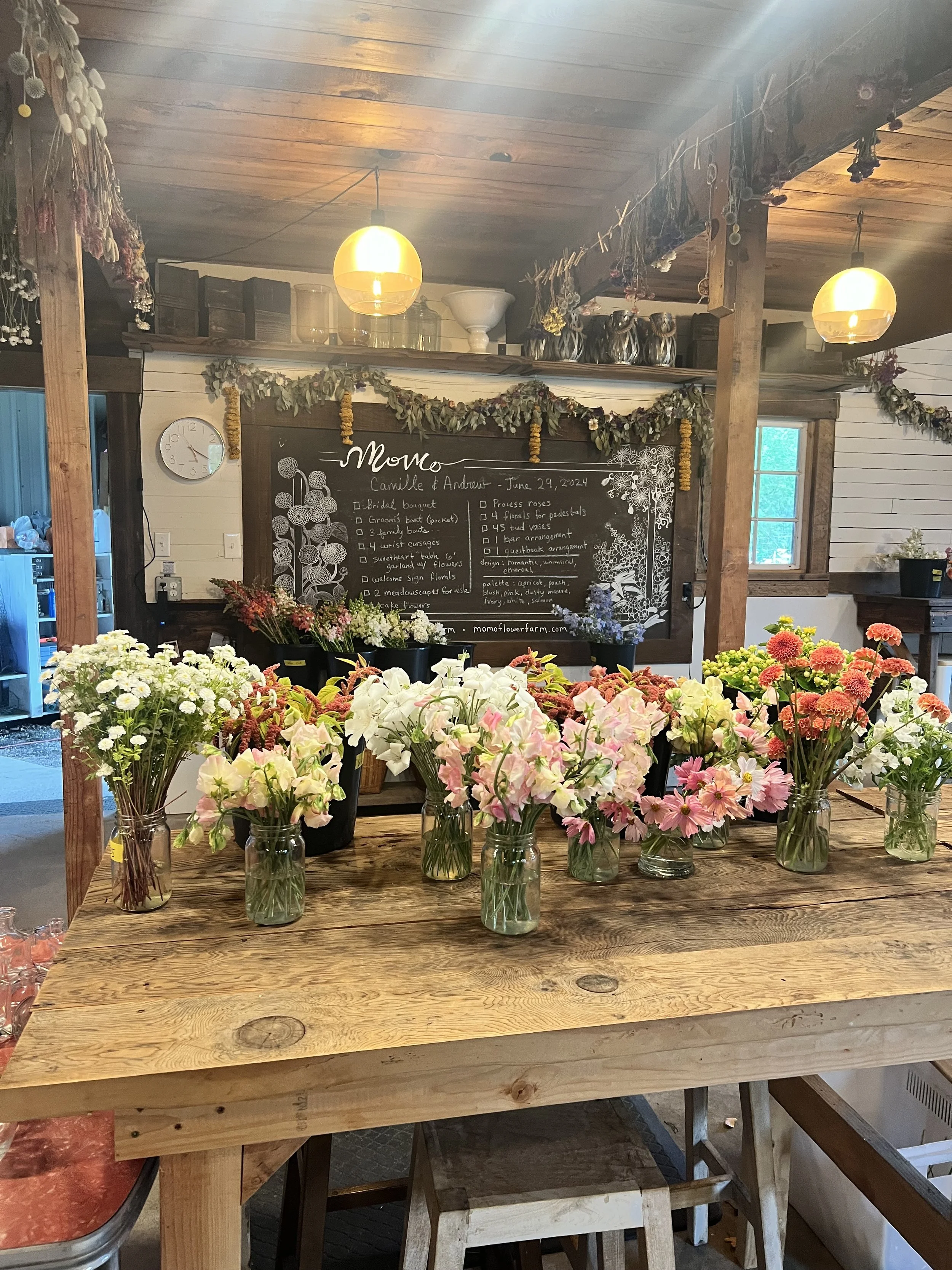 A wooden table with various colorful flowers in glass jars, inside a rustic, cozy shop or flower farm with a chalkboard menu in the background and warm lighting.