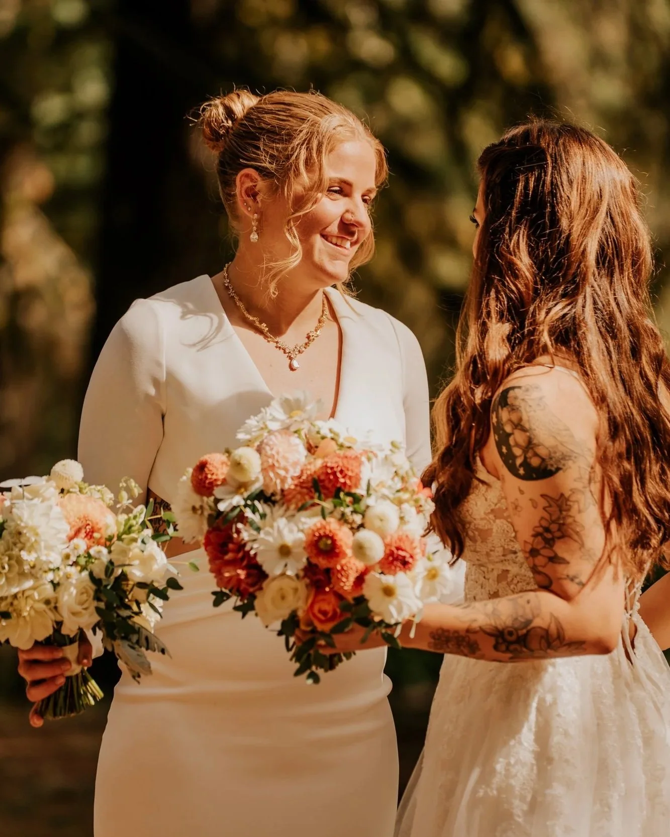Two women holding bouquets and smiling at each other during a wedding ceremony outdoors, with trees in the background.