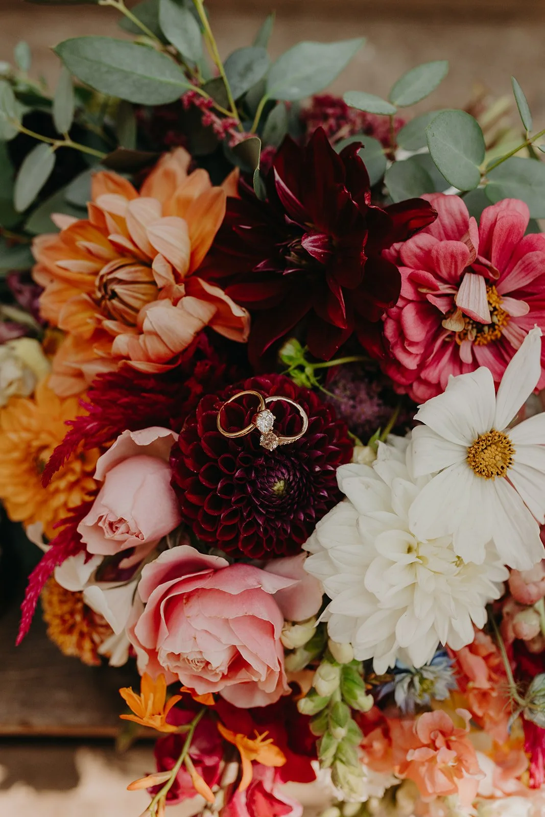 A colorful bouquet of various flowers, with a wedding ring placed on a dark red flower in the center.