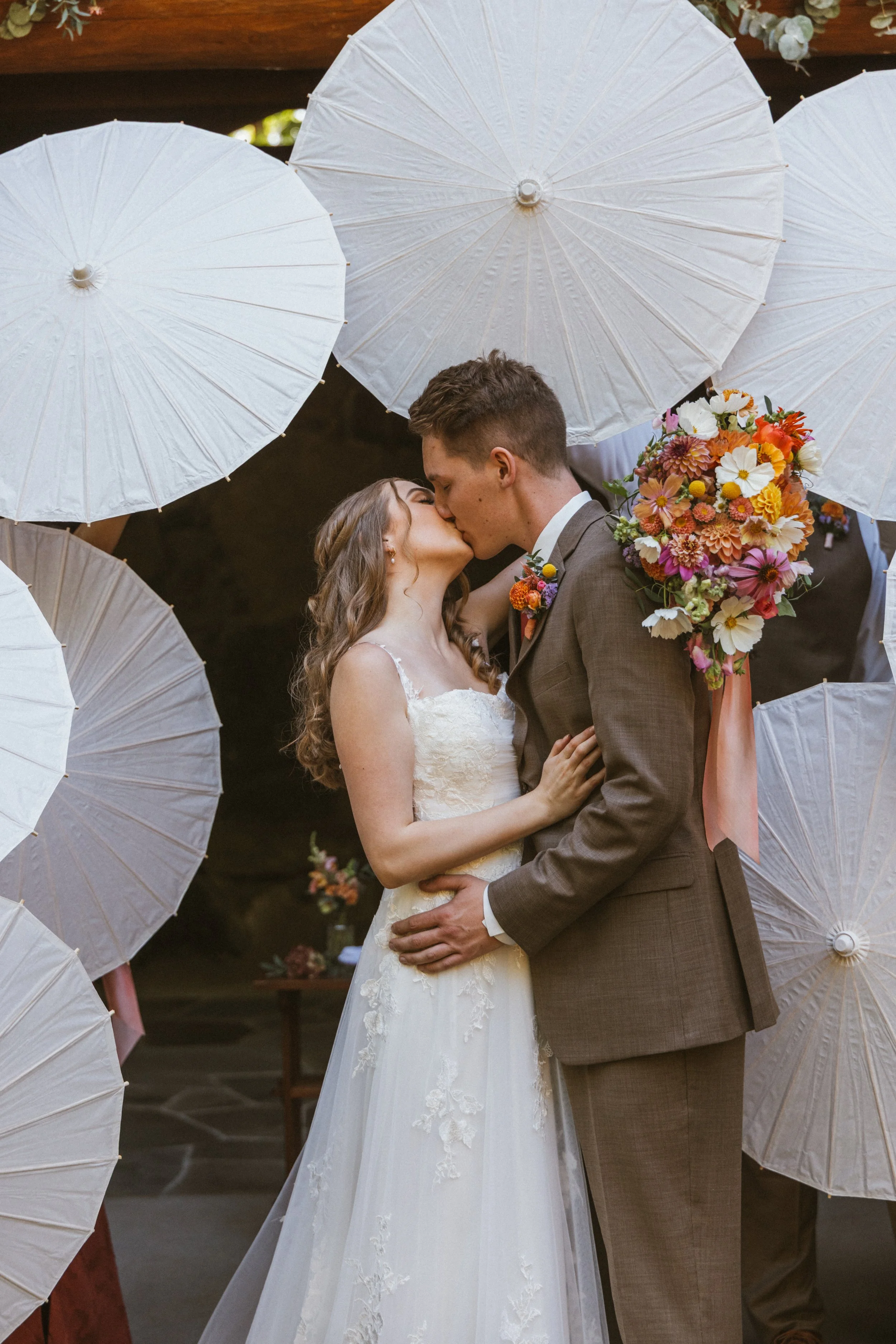A bride and groom sharing a kiss at their wedding surrounded by white umbrellas and a bouquet of colorful flowers.

Photo by Ivory Creek Photography
Venue: Summit Grove Lodge in Ridgefield, WA