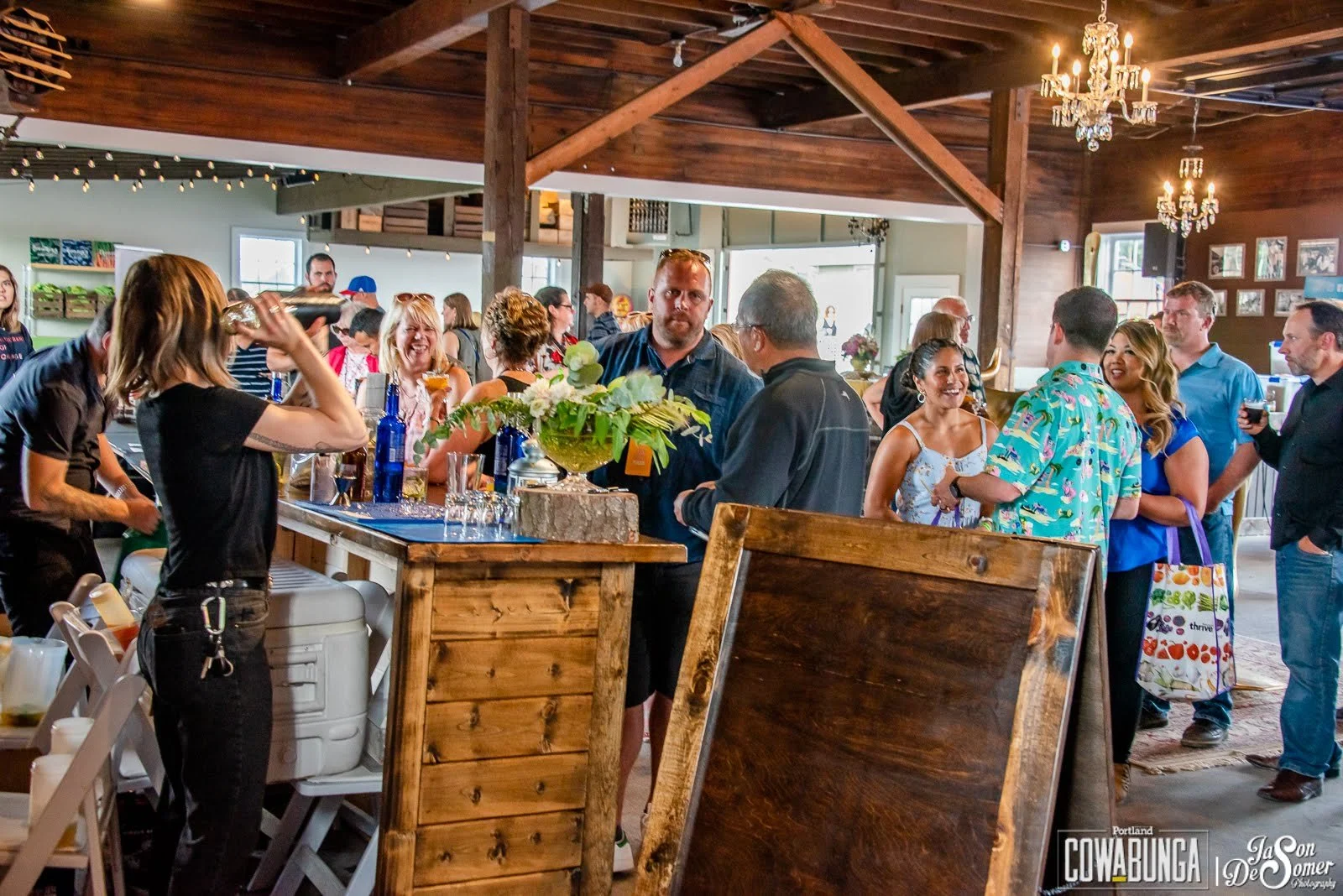 People socializing at a bar inside a rustic venue with wooden beams and chandeliers, some holding drinks and chatting.