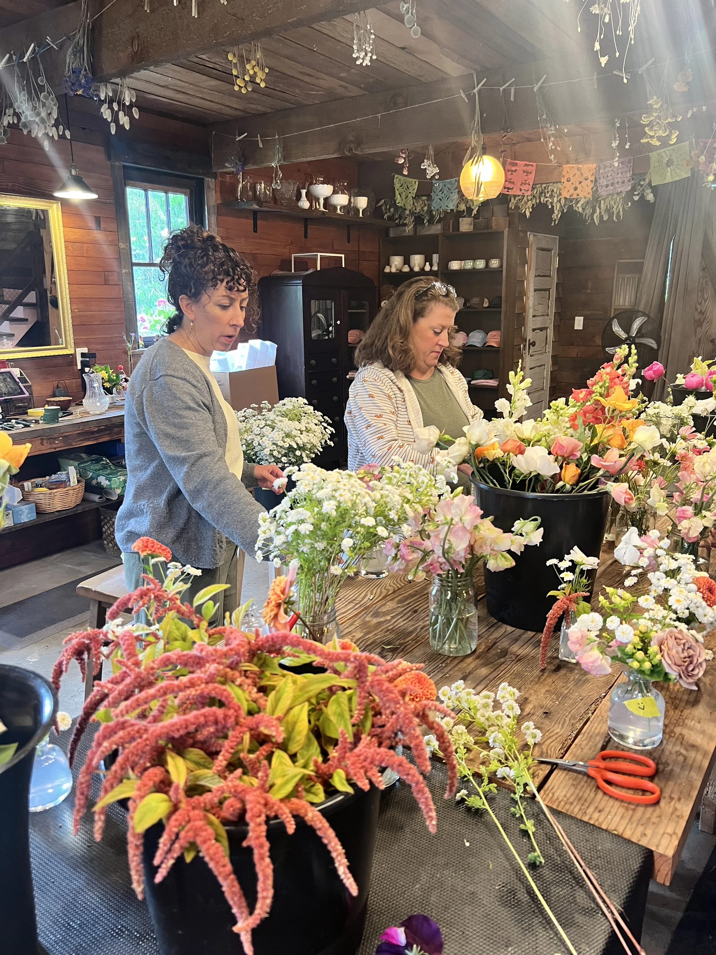 Two women arranging flowers in a rustic indoor space with wooden walls and ceiling, decorated with dried flowers and colorful papel picado banners.