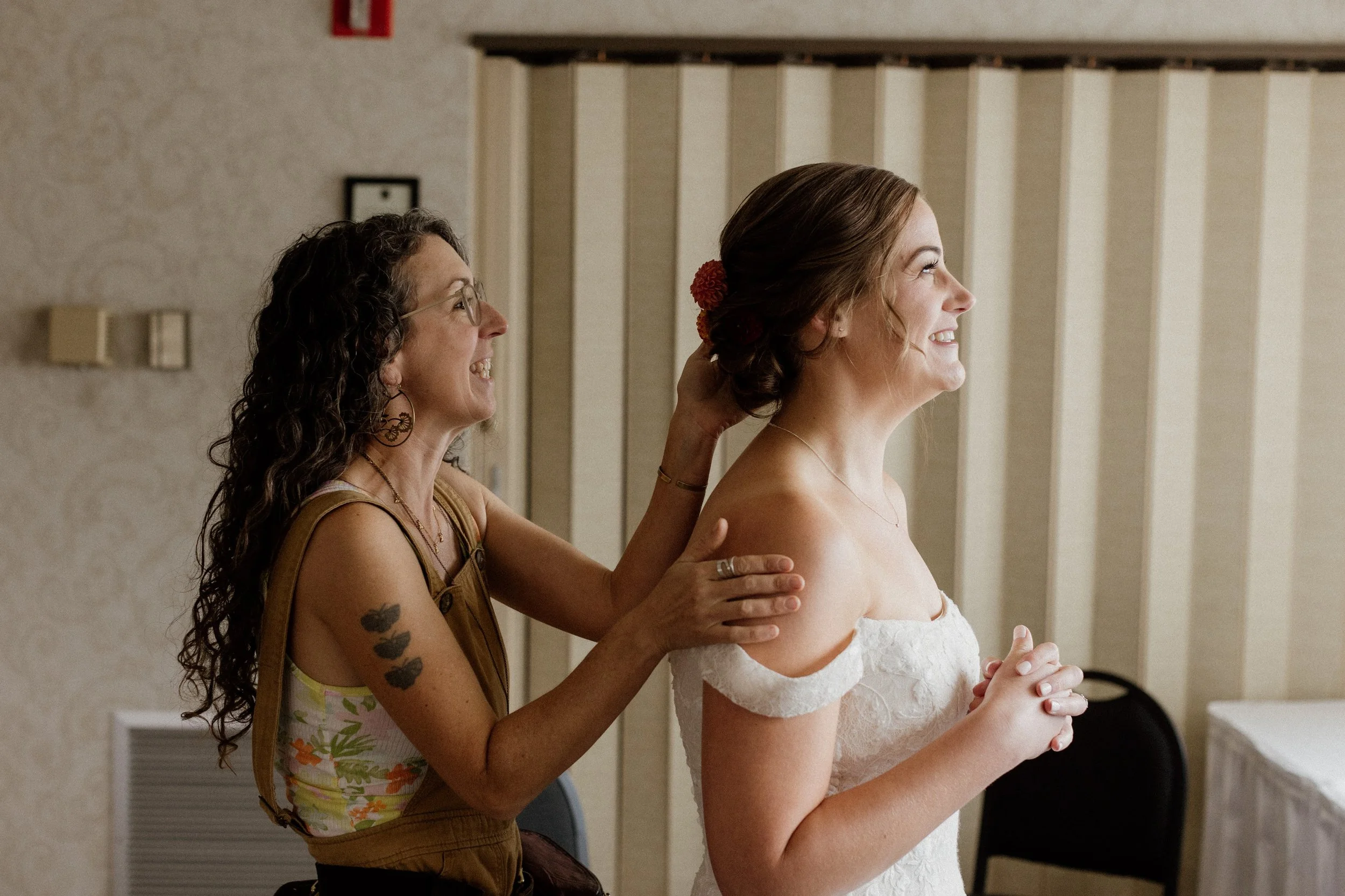A woman in a wedding dress smiling as another woman adjusts her hair in a room with patterned wallpaper. Photo by Kelly is Wild