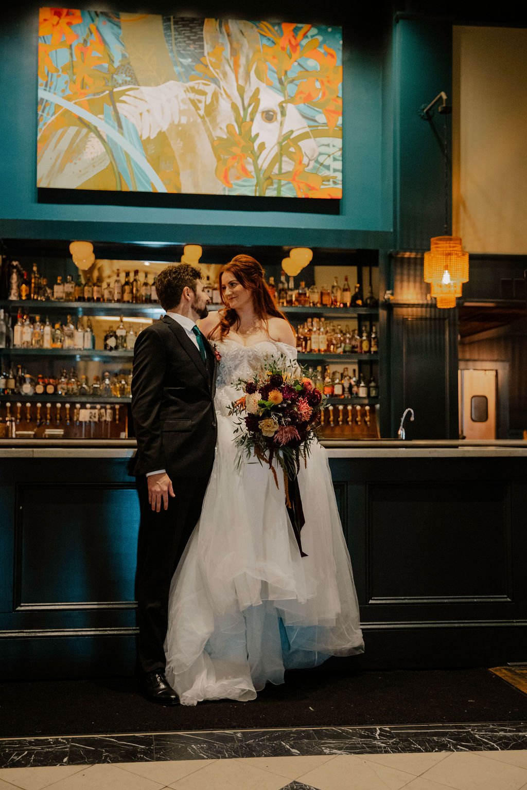 A newlywed couple in wedding attire standing close together at a bar, with the bride holding a large bouquet of flowers. Behind them is a well-stocked bar with bottles and taps, and a colorful artwork on the wall.

Photo by Ronny & Rene
Venue: Sentin
