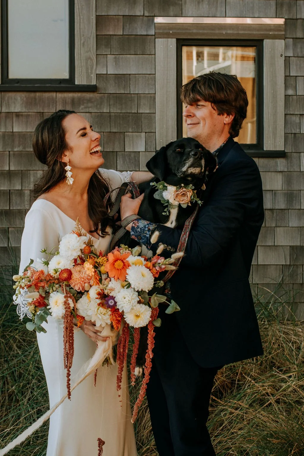 A bride and groom celebrating their wedding outdoors with their dog, who is wearing a floral collar, and the bride is holding a colorful bouquet.
