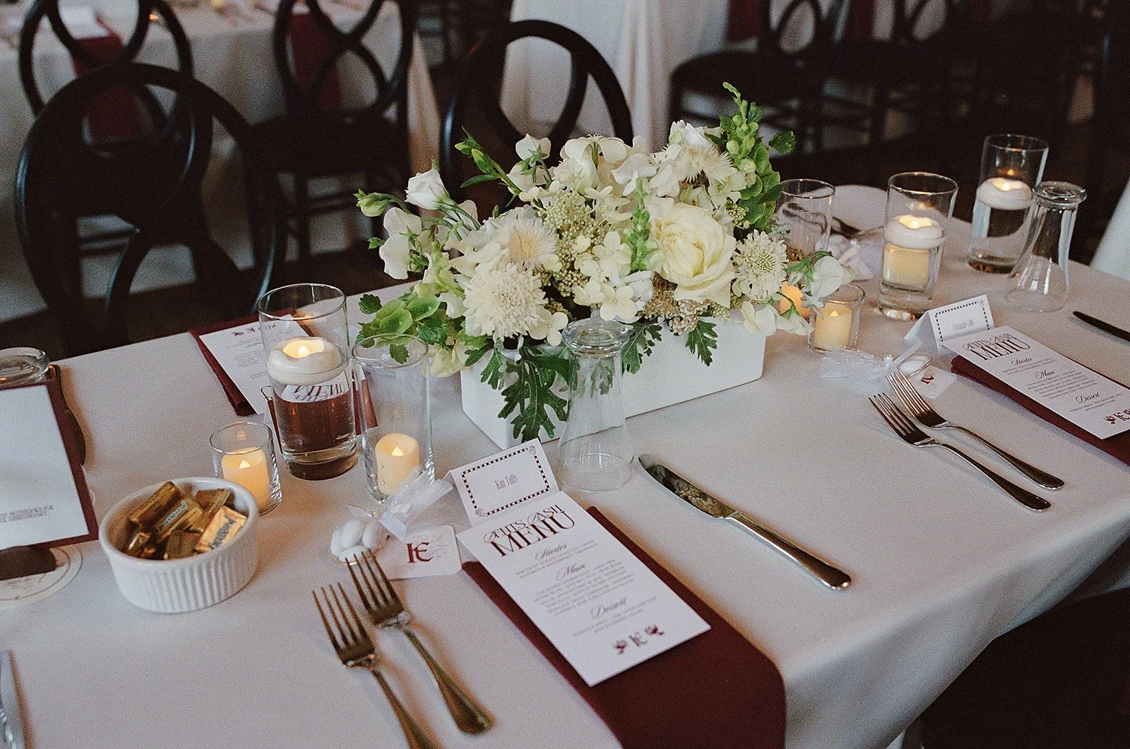 A formal dining table decorated with a white tablecloth, a centerpiece of white flowers, and small candles in glass holders. The table settings include forks, knives, and menus with maroon accents. There are empty chairs with dark frames around the t