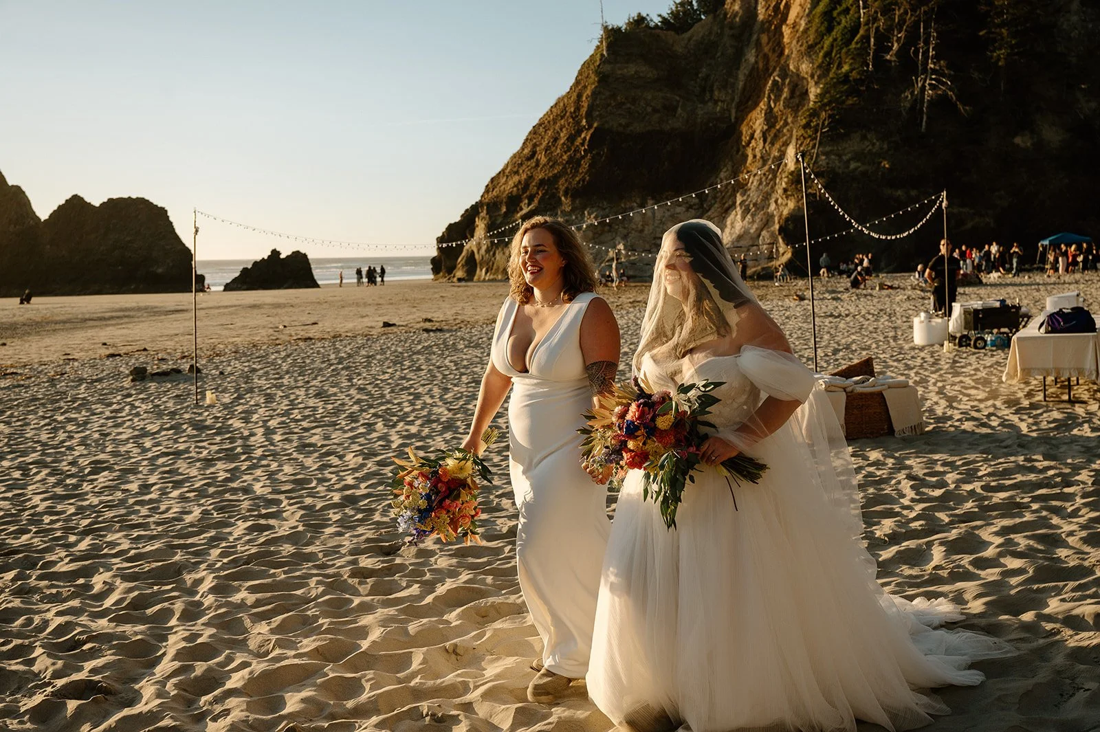 Two women in wedding dresses walking on a sandy beach during sunset, holding bouquets, with rocky formations and other people in the background.

Photo by Forgette Photo
Venue: the beautiful Oregon Coast