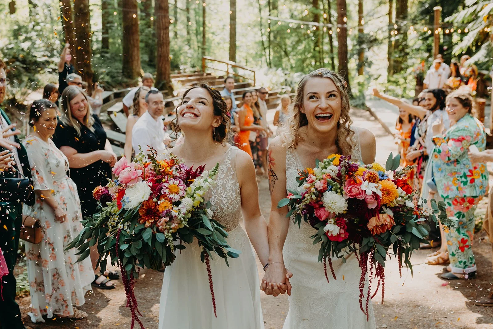 Two women in wedding dresses holding hands and carrying large bouquets of colorful flowers, smiling as they walk through a forested outdoor wedding ceremony with guests celebrating around them.

Photo by Olivia Louise Photography
Venue: Camp Cedarbro