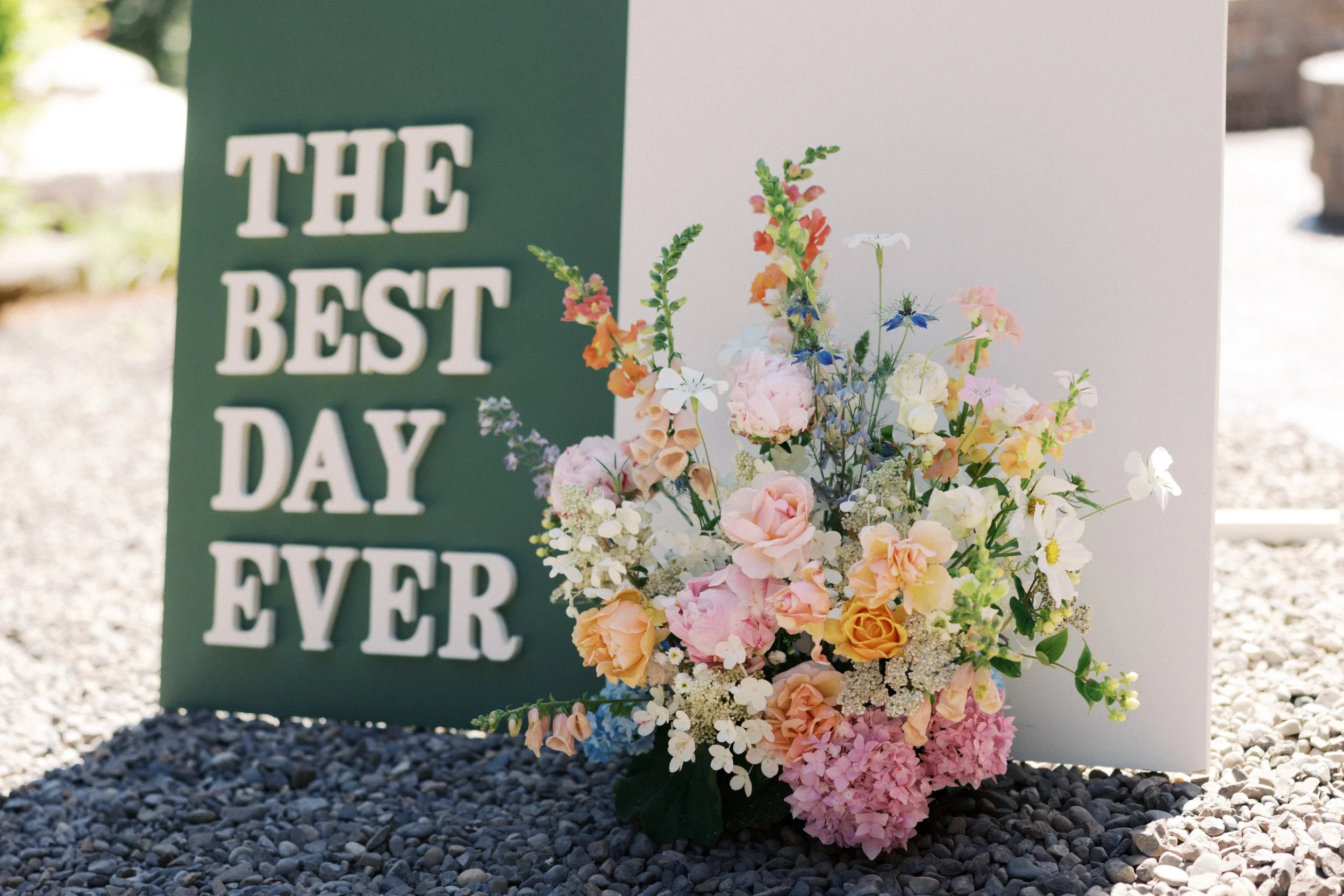 A wedding sign with the words 'The Best Day Ever' in white letters on a dark green background, adorned with a large bouquet of pink, white, and orange, and blue flowers.

Photo by Here Today Photography
Venue: Rocky Hill Weddings & Events in White Sa
