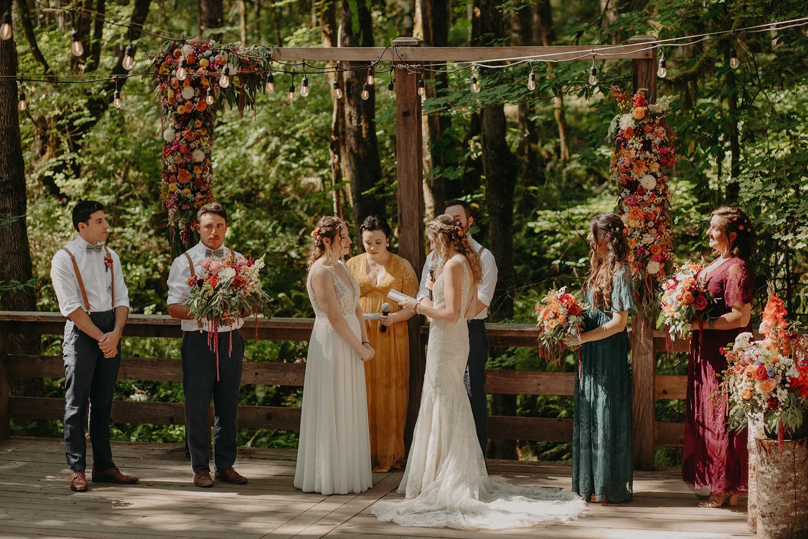 A wedding ceremony outdoors on a wooden deck, with two brides exchanging vows and holding hands, surrounded by six bridesmaids and groomsmen holding bouquets and standing in front of a floral arch, with lush greenery in the background.