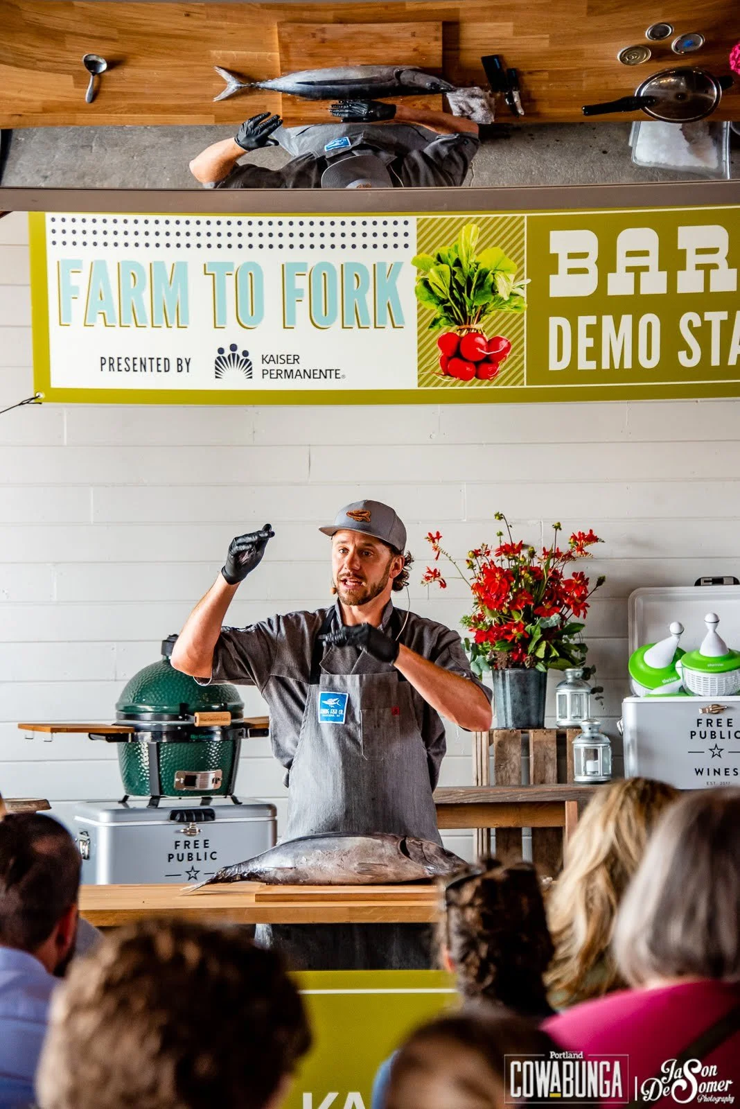 A man giving a fish cooking demonstration to an audience at a farm-to-fork bar demo stall, with a large fish on the table, a green grill in the background, and a sign reading 'Farm to Fork' and 'Bar Demo Stall'. Photo by Whatever Photo PDX