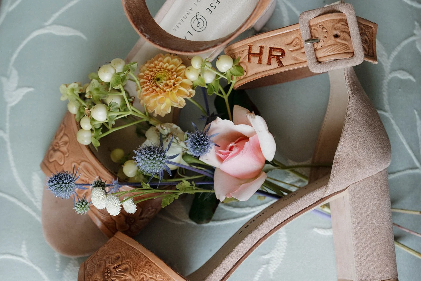 A pair of beige high-heeled shoes with floral decorations inside, including pink roses, blue thistles, white berries, a yellow dahlia, and white daisies, on a patterned fabric surface.
