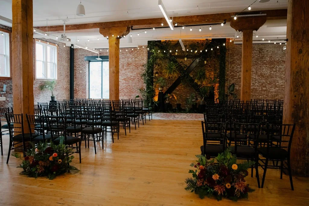 Interior of a rustic wedding or event venue with string lights, wooden beams, black chairs arranged in rows, exposed brick walls, large windows, and decorative floral arrangements on the floor.

Photo by Ronny & Rene
Venue: Ecotrust Events Irving Str