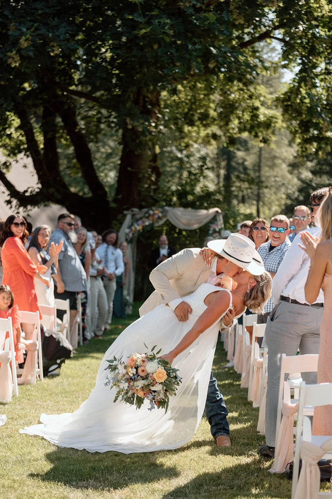 A couple shares a kiss at their outdoor wedding ceremony, with the bride holding a bouquet of flowers and the groom wearing a hat, while guests look on and smile.