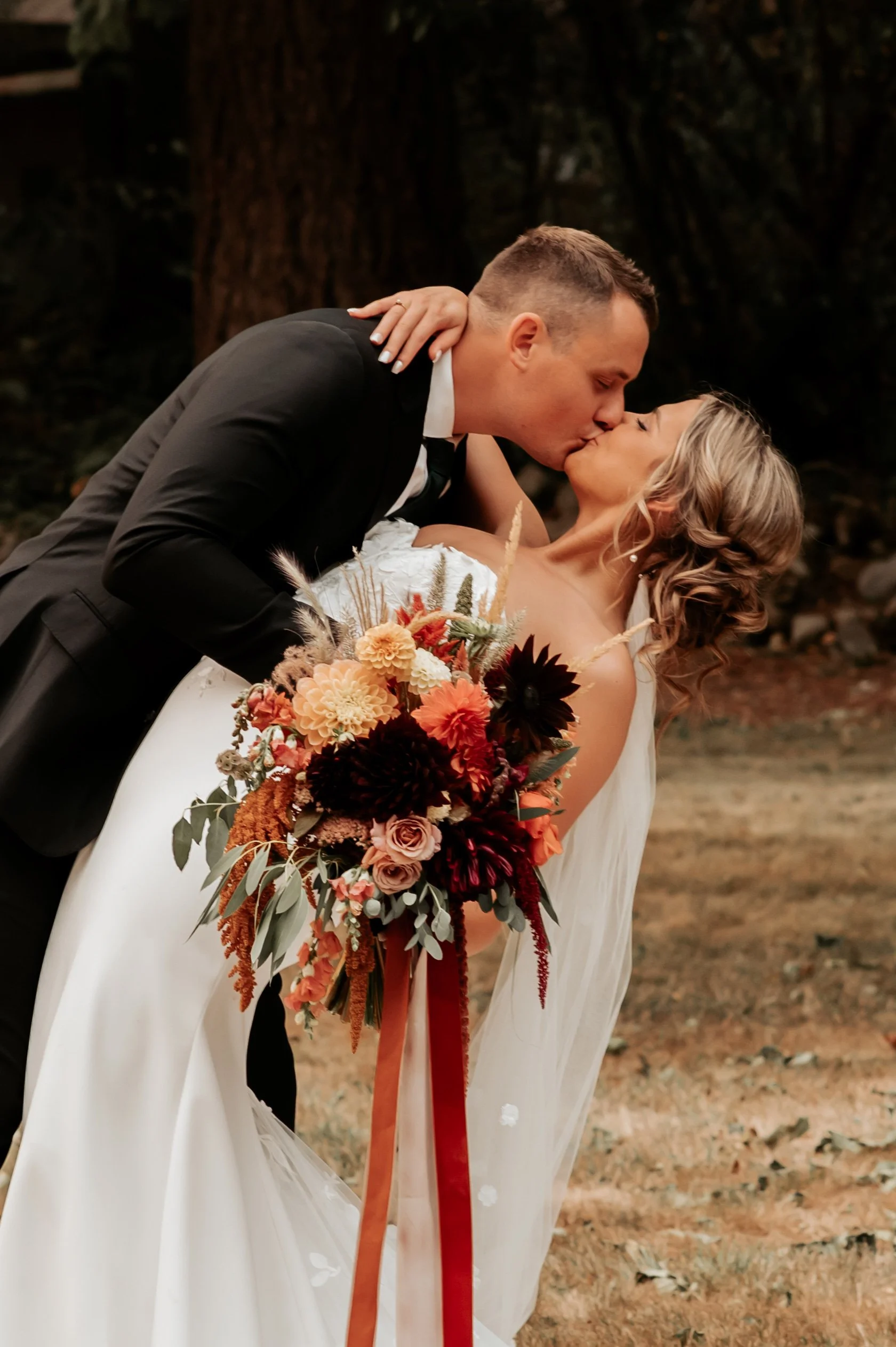 A groom in a black tuxedo kisses a bride in a white wedding dress outdoors, with a large bouquet of orange, peach, and burgundy flowers. Photo by Ashley Wilfong Photography