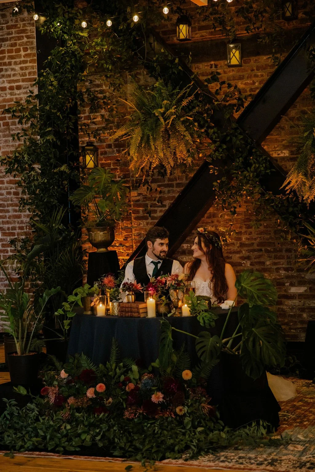 A couple dressed in wedding attire sitting at a sweetheart table decorated with candles, flowers, and greenery, surrounded by lush plants and a brick wall backdrop with hanging lanterns.

Photo by Ronny & Rene
Venue: Ecotrust Events Irving Street Stu
