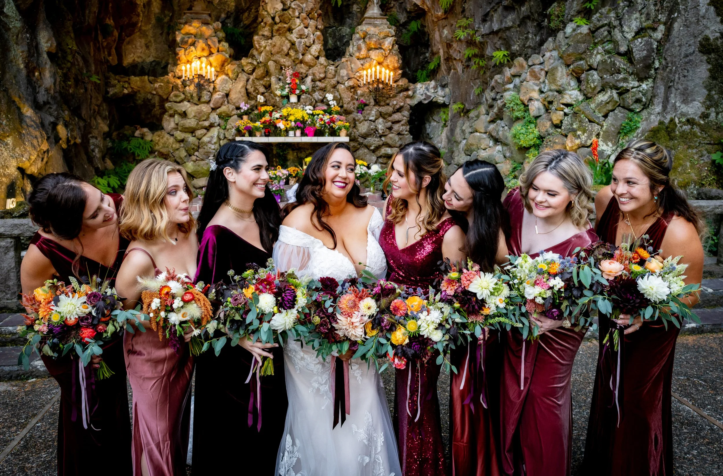 Bride and seven bridesmaids holding bouquets, standing in front of a stone wall with flowers and candles, at a wedding celebration.