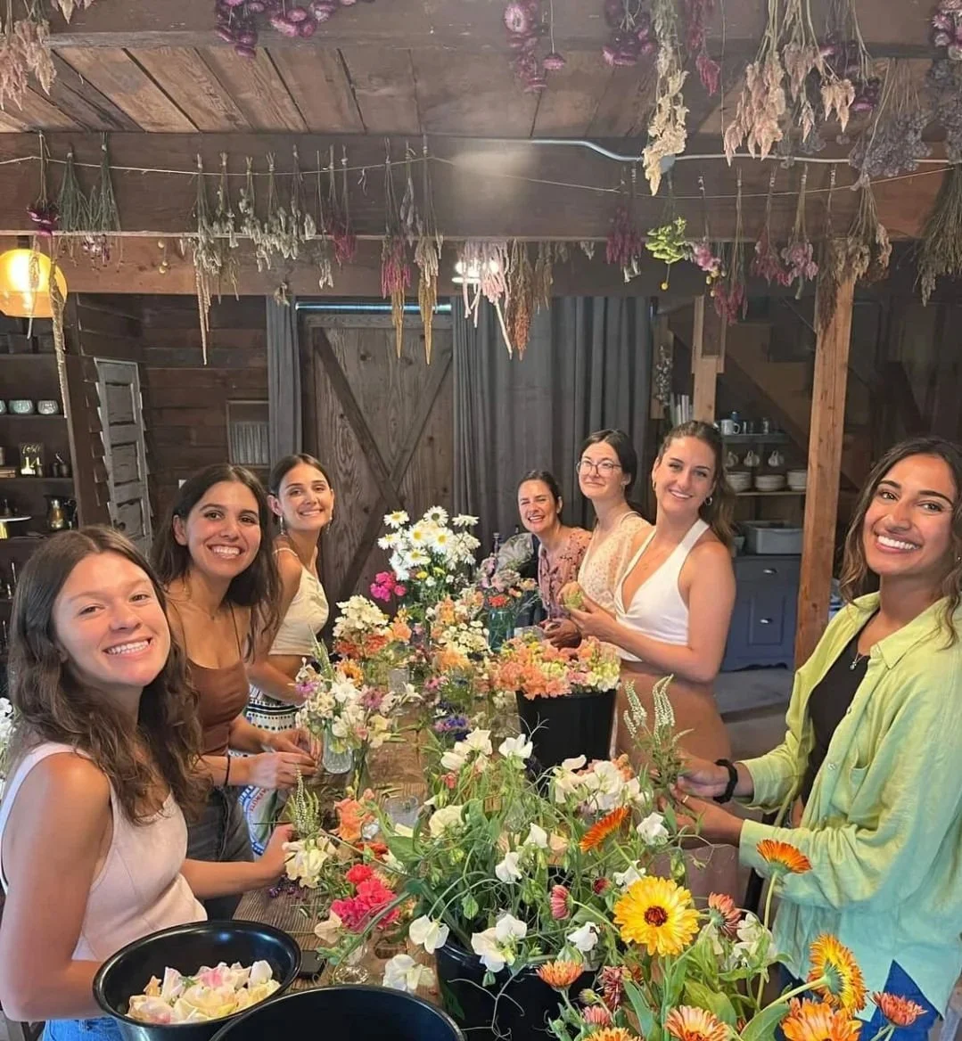 A group of seven women gathered around a table filled with colorful flowers in a rustic wooden room, smiling and enjoying a flower arranging activity.