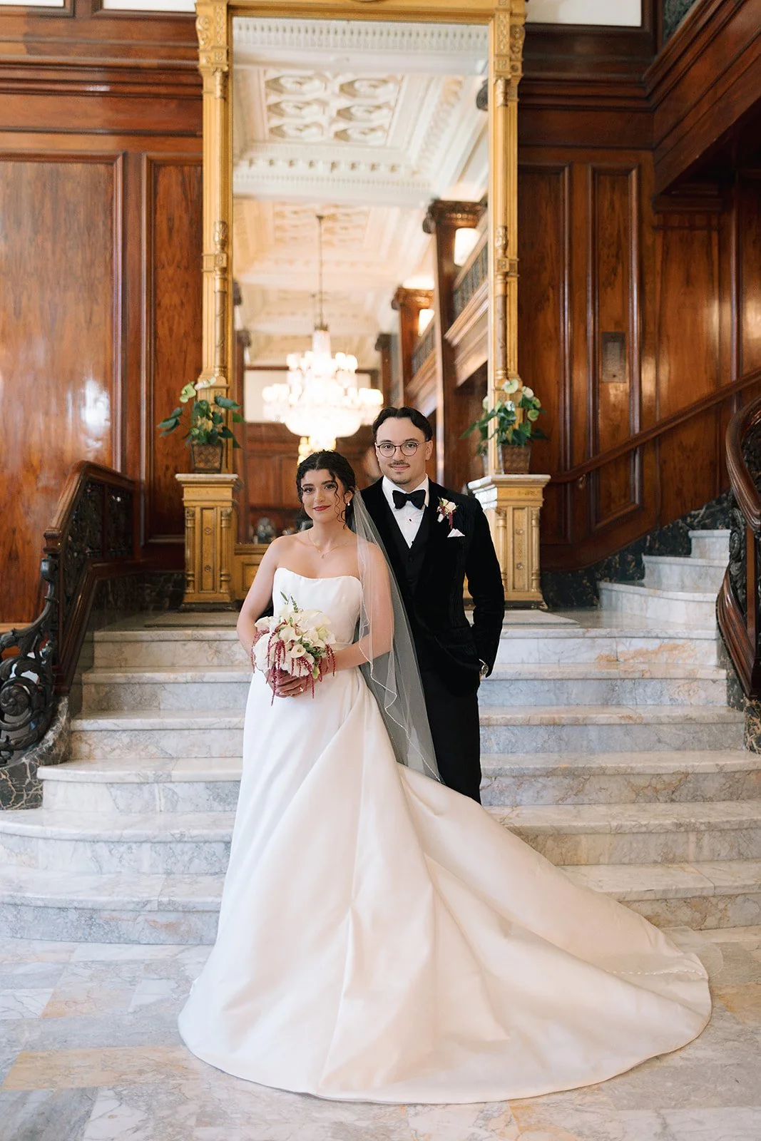 A bride in a white dress holding a bridal bouquet stands with a groom in a black suit in the grand hotel lobby of the Benson Hotel in Downtown Portland.

Photo by Mercedes Lou Photography
