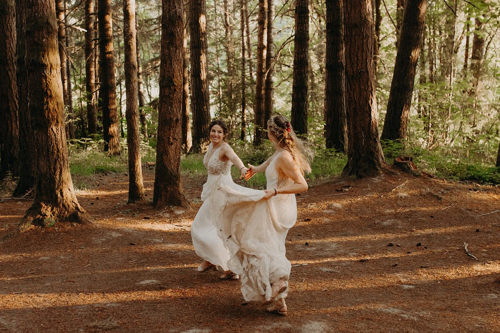 Two women in wedding dresses dancing in a forest with tall trees and sunlight filtering through.