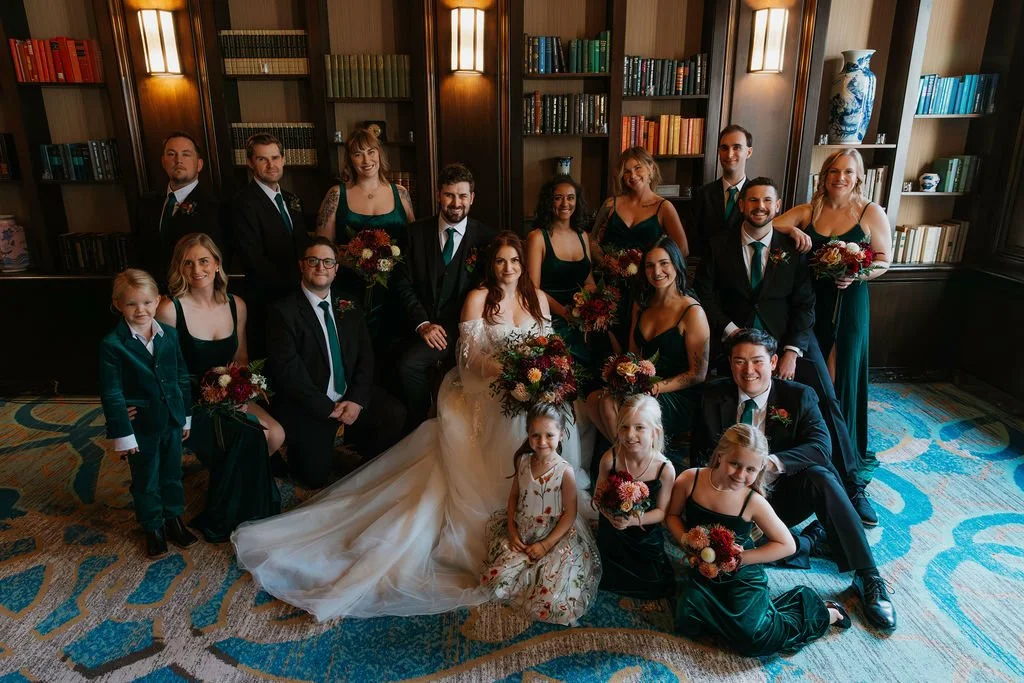 A wedding party group photo indoors with people dressed in formal attire, with the bride in a white wedding dress sitting in the center surrounded by friends and family holding bouquets of flowers.

Photo by Ronny & Rene Photography
Venue: Sentinel H