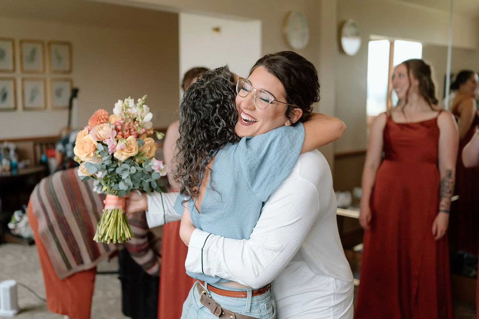 Two women hugging and smiling, one holding a bouquet of flowers, at a social gathering indoors.