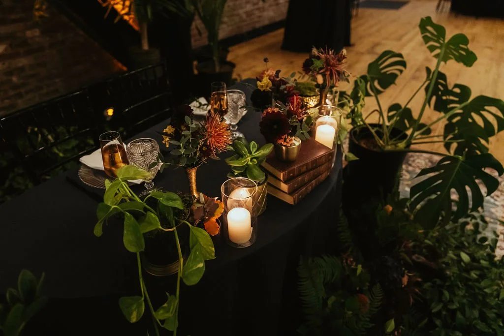 A round table set for a gathering with a black tablecloth, decorated with candles, flowers, and greenery, including potted plants and leafy branches, in a romantically lit indoor space.

Photo by Ronny & Rene Photography
Venue: EcoTrust Events Irving