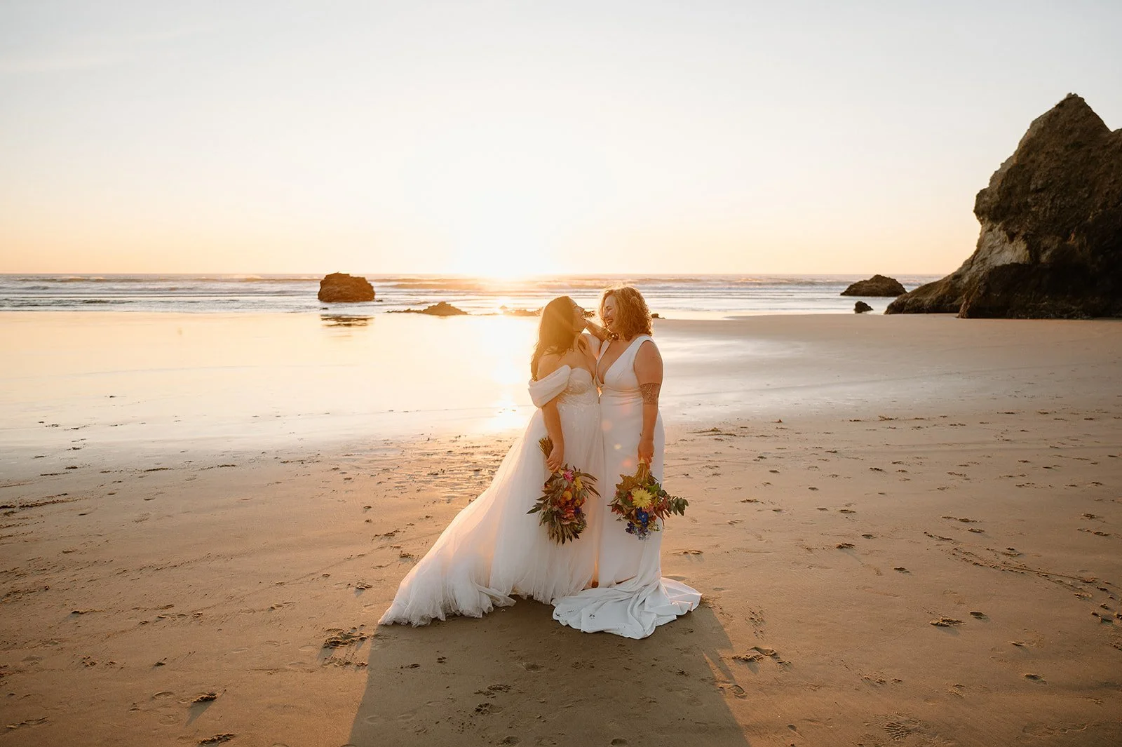 Two women in wedding dresses holding bouquets standing on a beach at sunset, with the ocean and rocks in the background.

Photo by Forgette Photo
Venue: the beautiful Oregon Coast