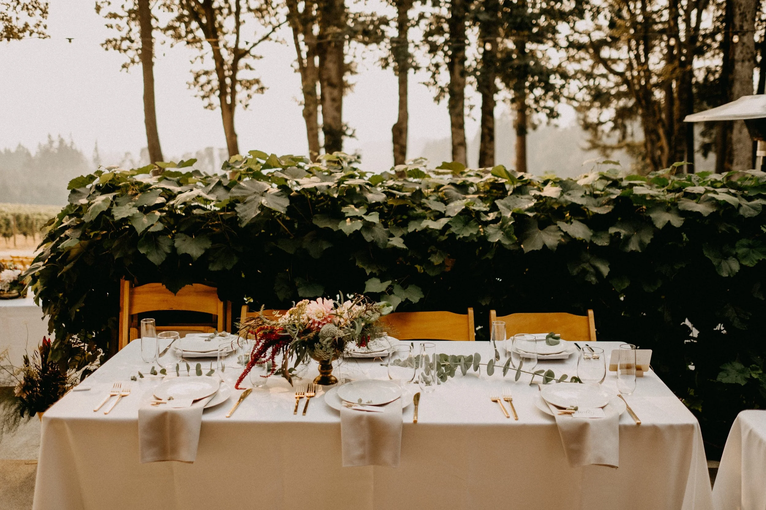 A rectangular dining table set for a meal with white tablecloth, gold utensils, white plates, clear glasses, and a floral centerpiece, outdoors in a garden with leafy bushes and trees in the background.