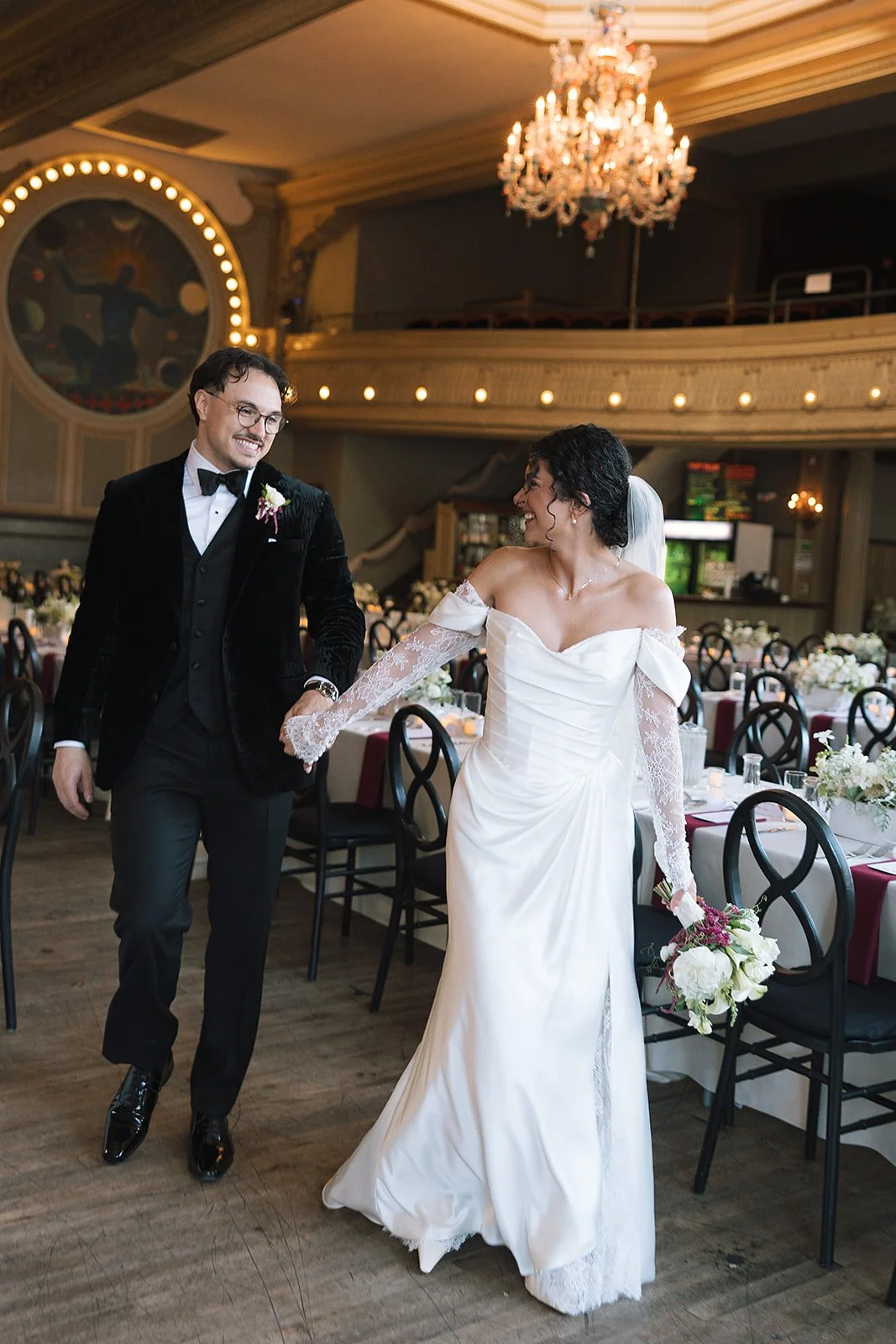 A bride and groom walking together in a wedding reception hall, holding hands and smiling at each other, with decorated tables and a large chandelier overhead.