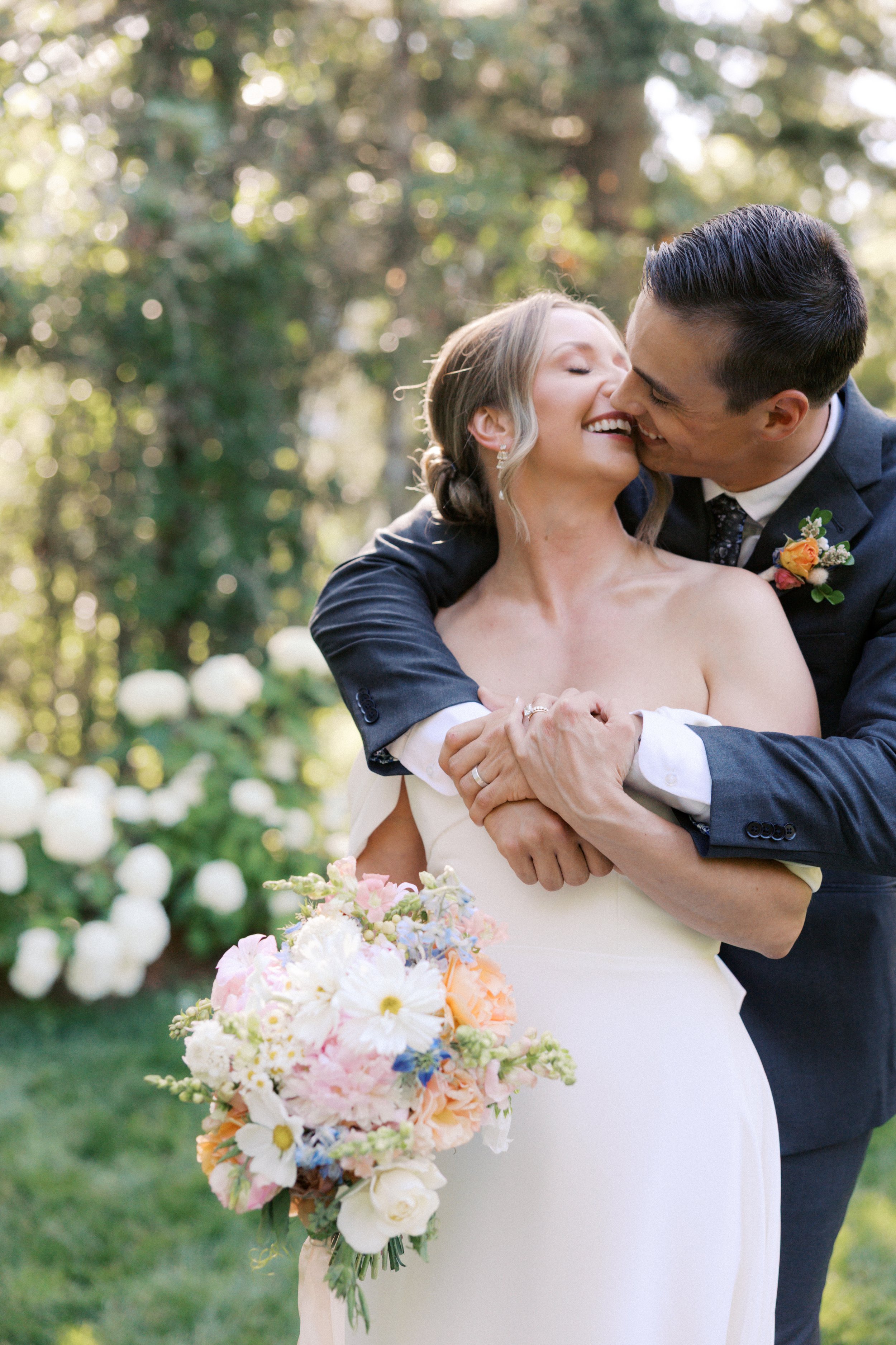 A newlywed couple shares a romantic hug outdoors, with trees and white flowers in the background. The bride wears a strapless white wedding dress and holds a bouquet of colorful flowers, while the groom, in a dark suit, leans in to kiss her.

Photo b