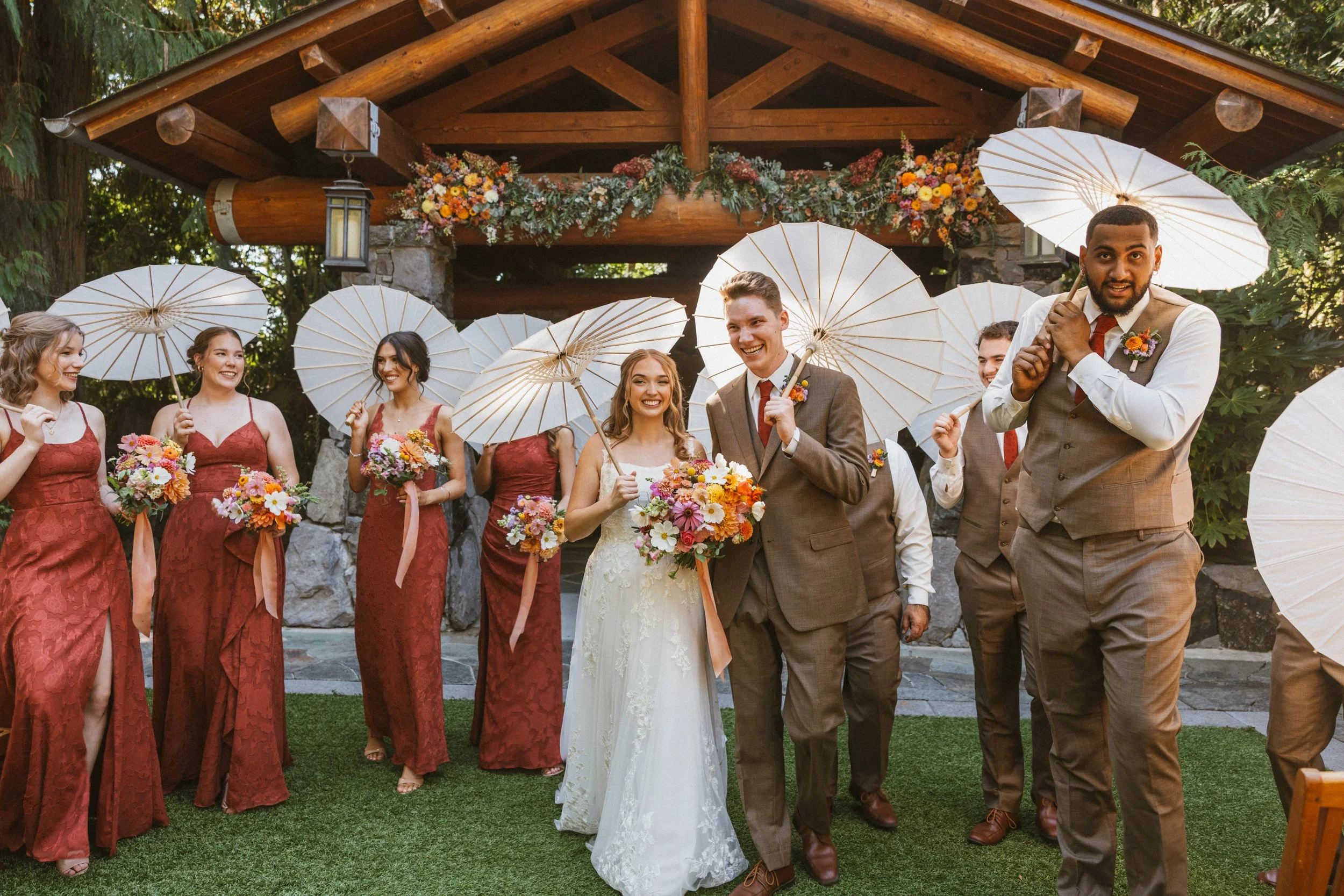 A wedding celebration with the bride and groom walking arm in arm, surrounded by bridesmaids and groomsmen holding white parasols, in an outdoor setting with a wooden arch decorated with flowers in the background.

Photo by Ivory Creek Photography
Ve